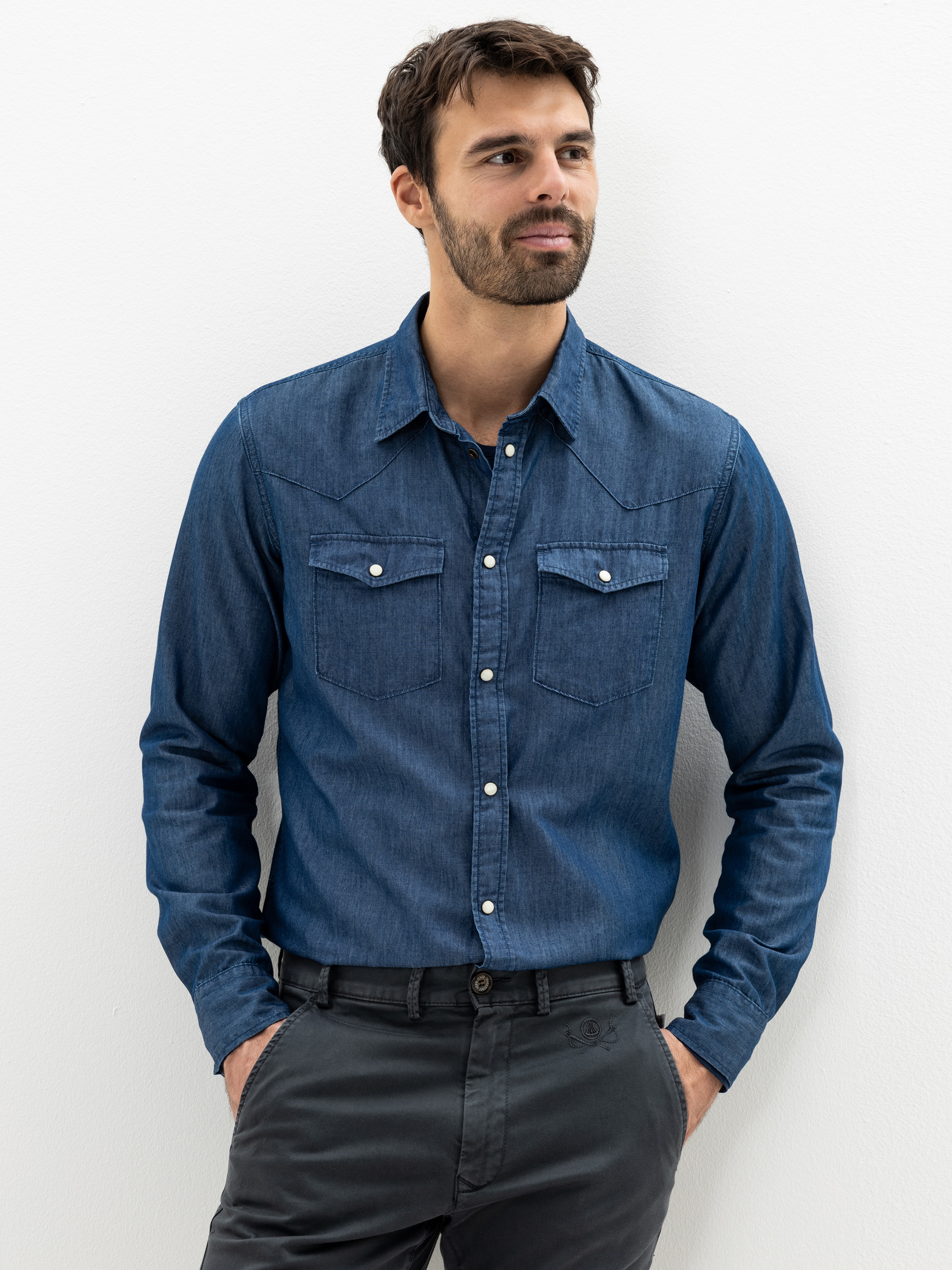 Ein Mann mit kurzen dunklen Haaren und einem Bart steht vor einem weißen Hintergrund. Er trägt ein blaues Jeanshemd zum Aufknöpfen mit Vordertaschen und eine dunkelgraue Hose. Er hat die Hände in den Taschen und blickt leicht zur Seite.