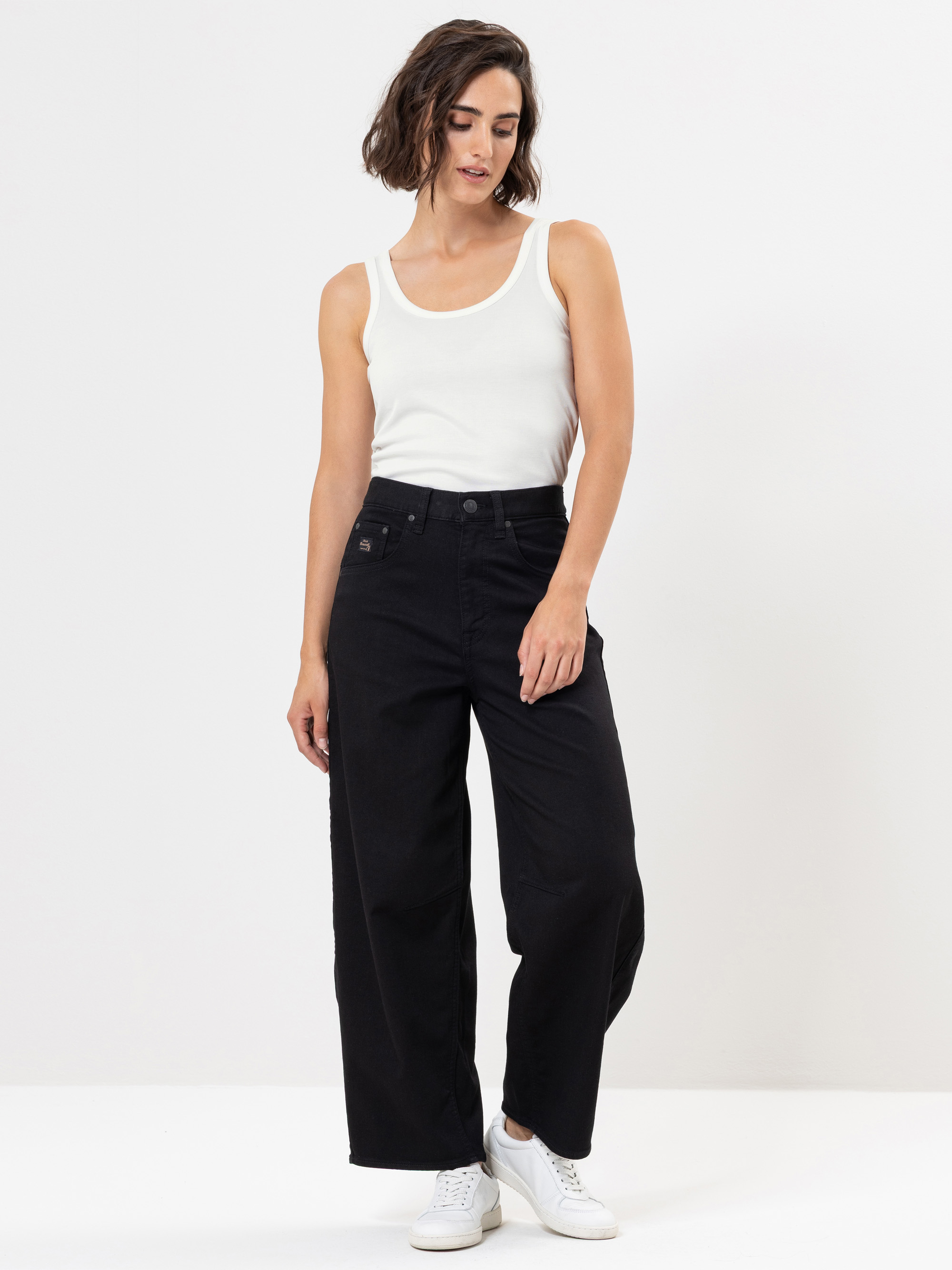 Eine Frau steht vor einem weißen Hintergrund und trägt ein weißes ärmelloses Oberteil, eine hochgeschlossene schwarze Hose mit weitem Bein und weiße Turnschuhe. Sie hat kurzes, gewelltes braunes Haar und blickt nach unten, wobei eine Hand auf ihrem O