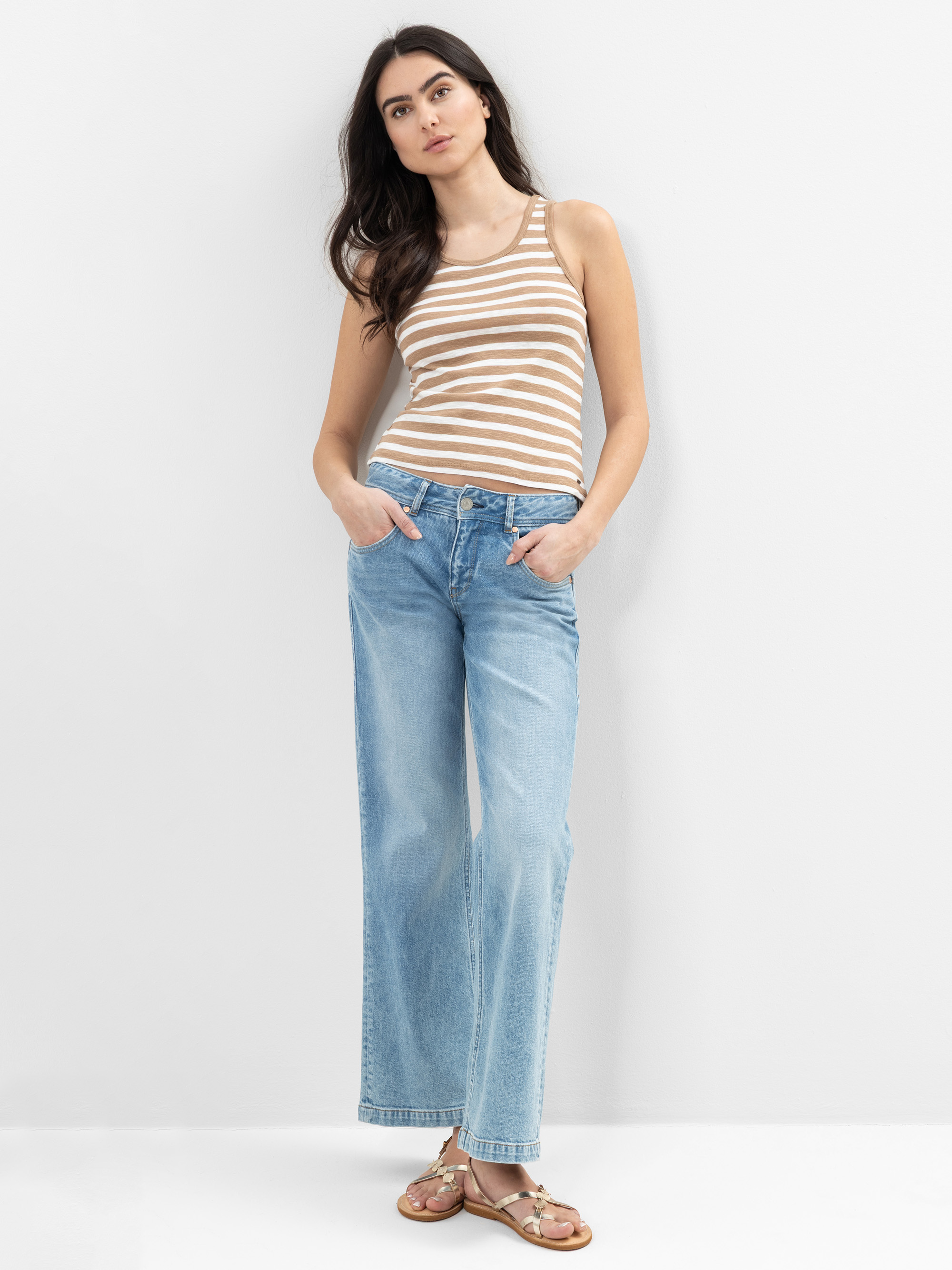 Eine Frau steht vor einer weißen Wand und trägt ein beige-weiß gestreiftes Tank-Top, hellblaue Jeans mit weitem Bein und Sandalen. Sie hat langes dunkles Haar und ihre Hände stecken in den Taschen.