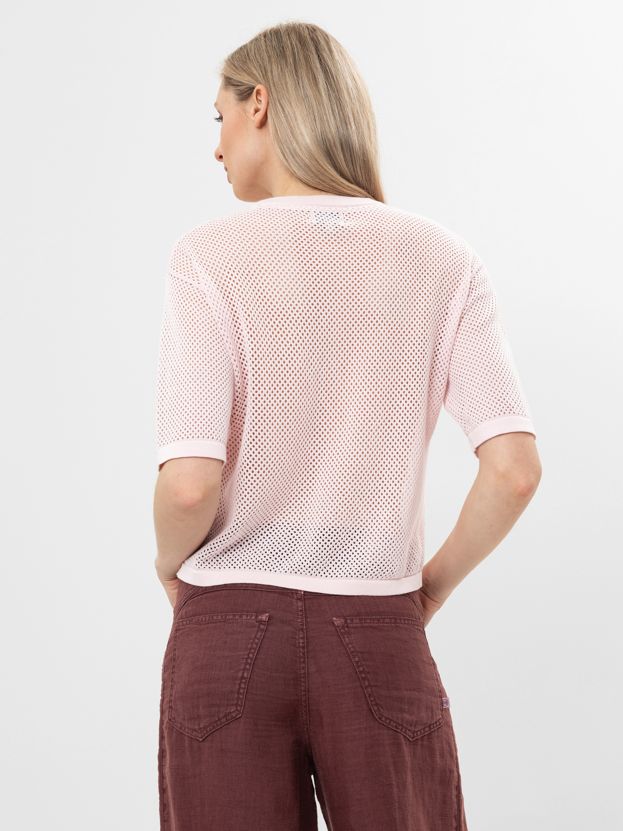 Eine Frau mit langen blonden Haaren, von hinten gesehen, trägt ein hellrosa, kurzärmeliges Netzoberteil und eine kastanienbraune Hose mit hoher Taille. Sie steht vor einem schlichten weißen Hintergrund.