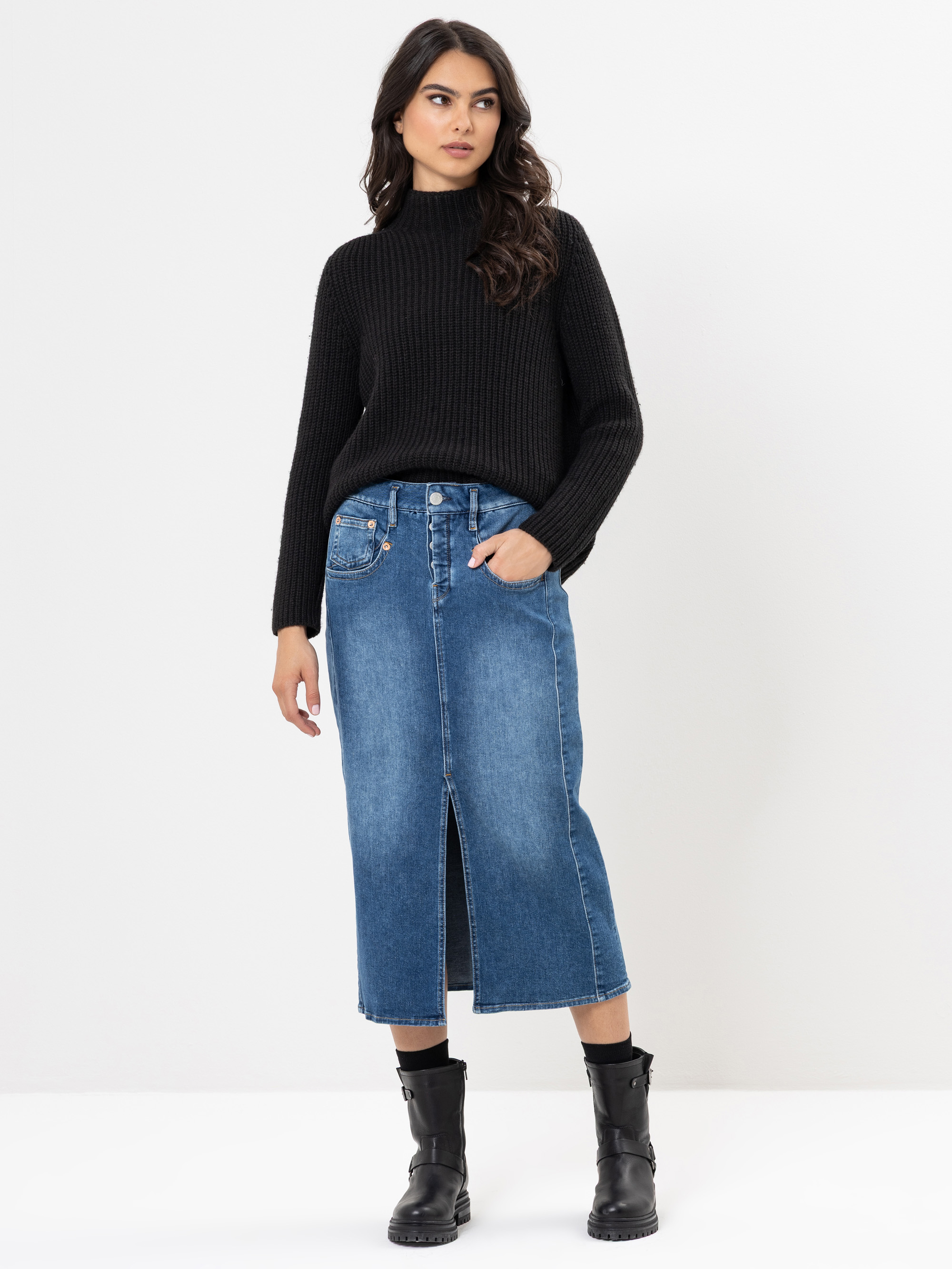 Eine Frau steht vor einem weißen Hintergrund und trägt einen schwarzen gerippten Pullover, einen blauen Jeans-Midirock mit einem vorderen Schlitz und schwarze Ankle Boots. Sie hat eine Hand in ihrer Rocktasche und blickt leicht zur Seite.