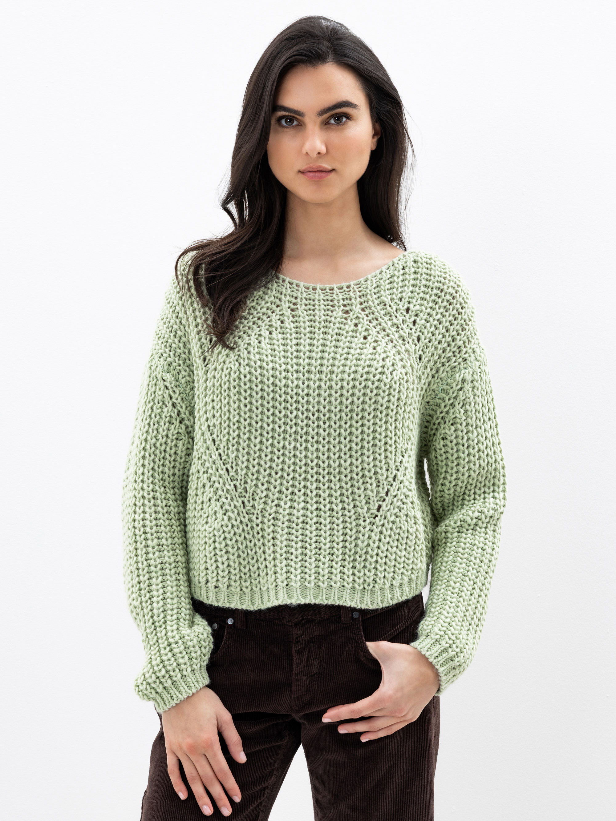 Eine Frau mit langen dunklen Haaren, die einen hellgrünen Strickpullover und eine dunkelbraune Hose trägt, steht vor einem schlichten weißen Hintergrund und blickt mit neutraler Miene in die Kamera.