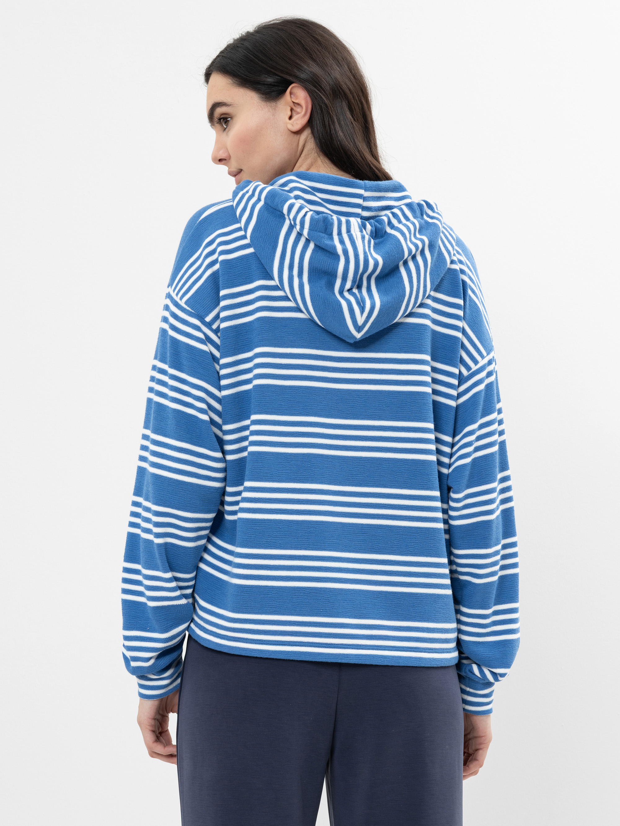Eine Frau steht in einem blau-weiß gestreiften Kapuzensweatshirt mit heruntergelassener Kapuze und einer weiten marineblauen Hose vor einem schlichten hellen Hintergrund.