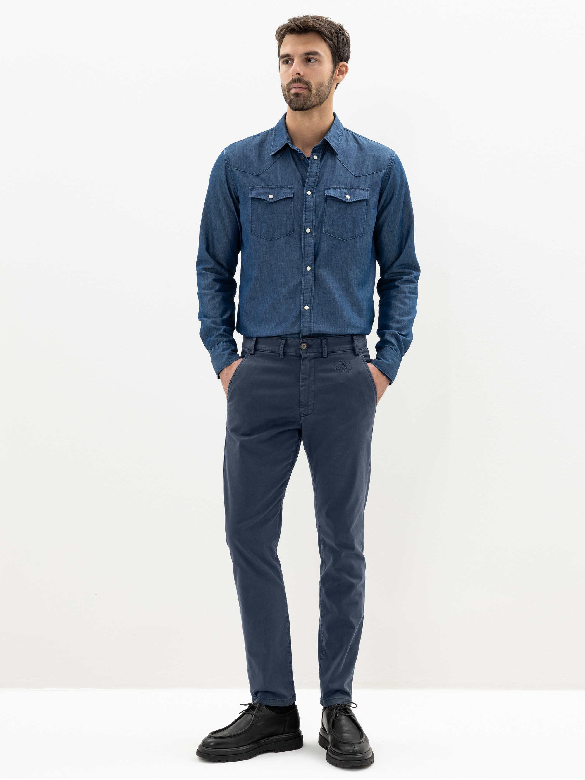 Ein Mann steht vor einem schlichten weißen Hintergrund und trägt ein blaues Hemd mit Knopfleiste und Vordertaschen, eine blaue Chinohose und schwarze Anzugschuhe. Er blickt nach vorne und hat die Hände in den Taschen.