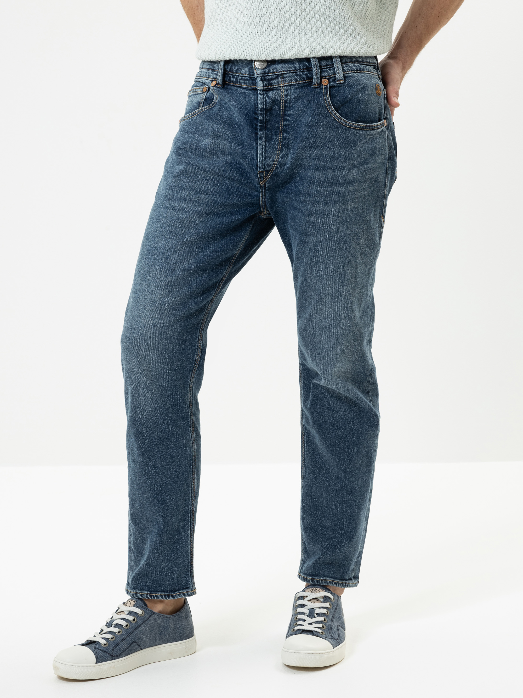 Eine Person in blauen Jeans und blauen Turnschuhen steht vor einem schlichten weißen Hintergrund und hat eine Hand in der Tasche. Ihr Oberkörper ist teilweise außerhalb des Bildausschnitts.