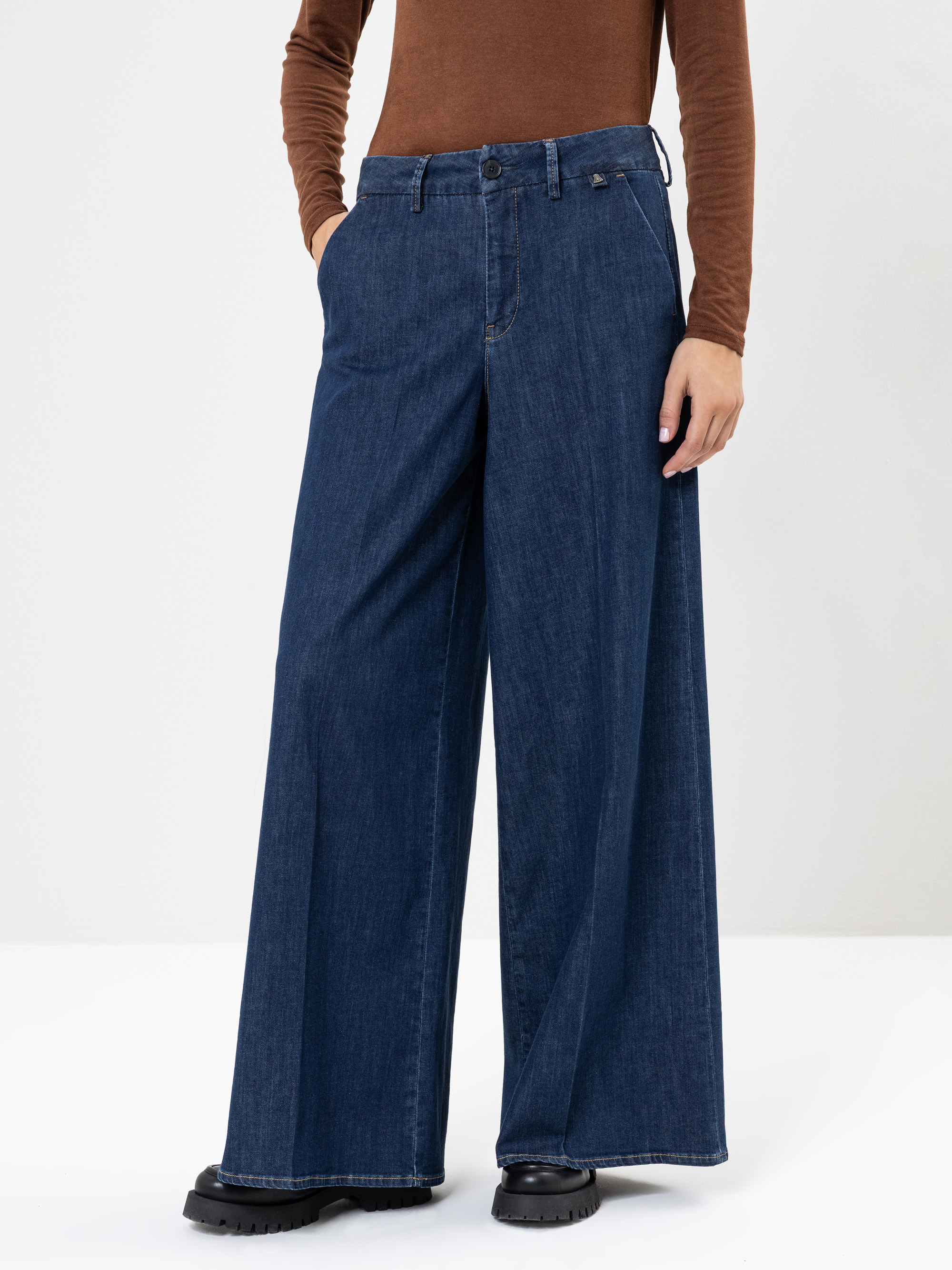 Eine Person, die ein braunes, langärmeliges Oberteil, eine weit geschnittene Jeans und schwarze Schuhe trägt, steht mit den Händen in den Taschen vor einem schlichten weißen Hintergrund. Ihr Kopf und ihre obere Brust sind nicht zu sehen.