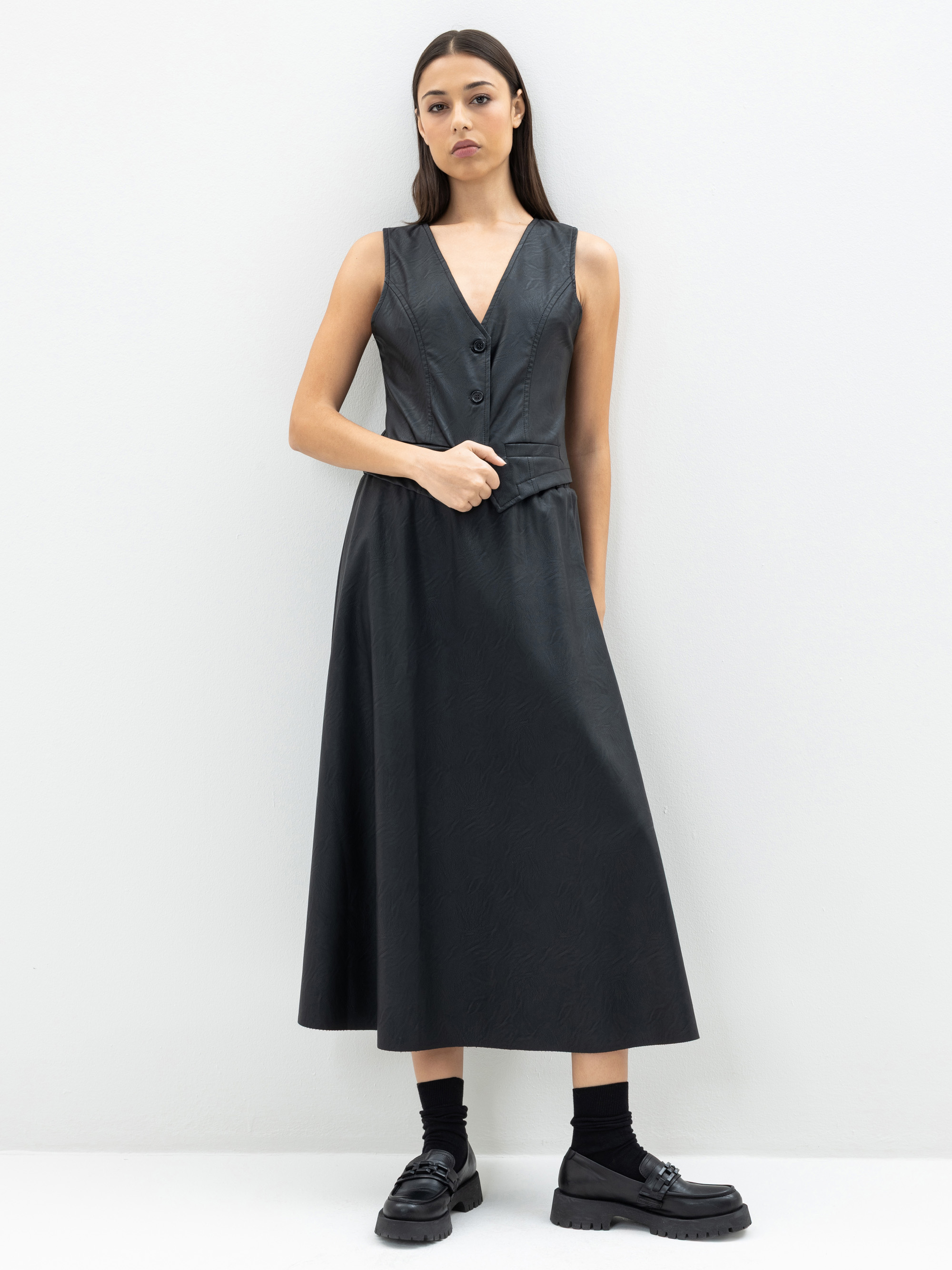 Eine Frau steht vor einem schlichten, hellen Hintergrund und trägt ein ärmelloses, schwarzes Kleid mit tiefem V-Ausschnitt und Knöpfen, schwarze Socken und klobige schwarze Schuhe. Sie hat langes, glattes Haar und einen neutralen Gesichtsausdruck.