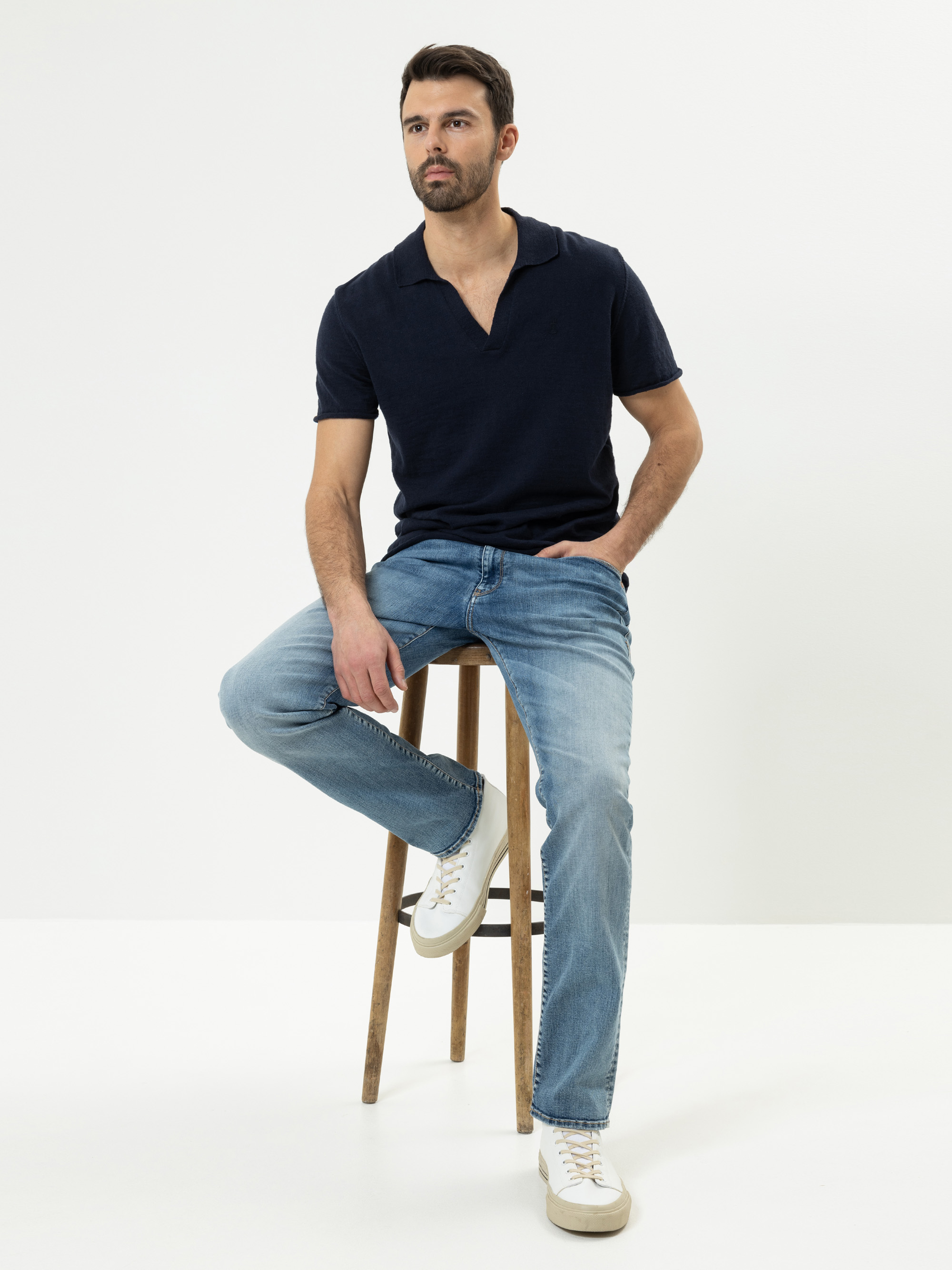 Ein Mann mit kurzen dunklen Haaren und einem Bart sitzt auf einem Holzhocker vor einem schlichten weißen Hintergrund. Er trägt ein marineblaues Polohemd, hellblaue Jeans und weiße Turnschuhe. Eine Hand steckt in seiner Hosentasche, und er schaut leicht