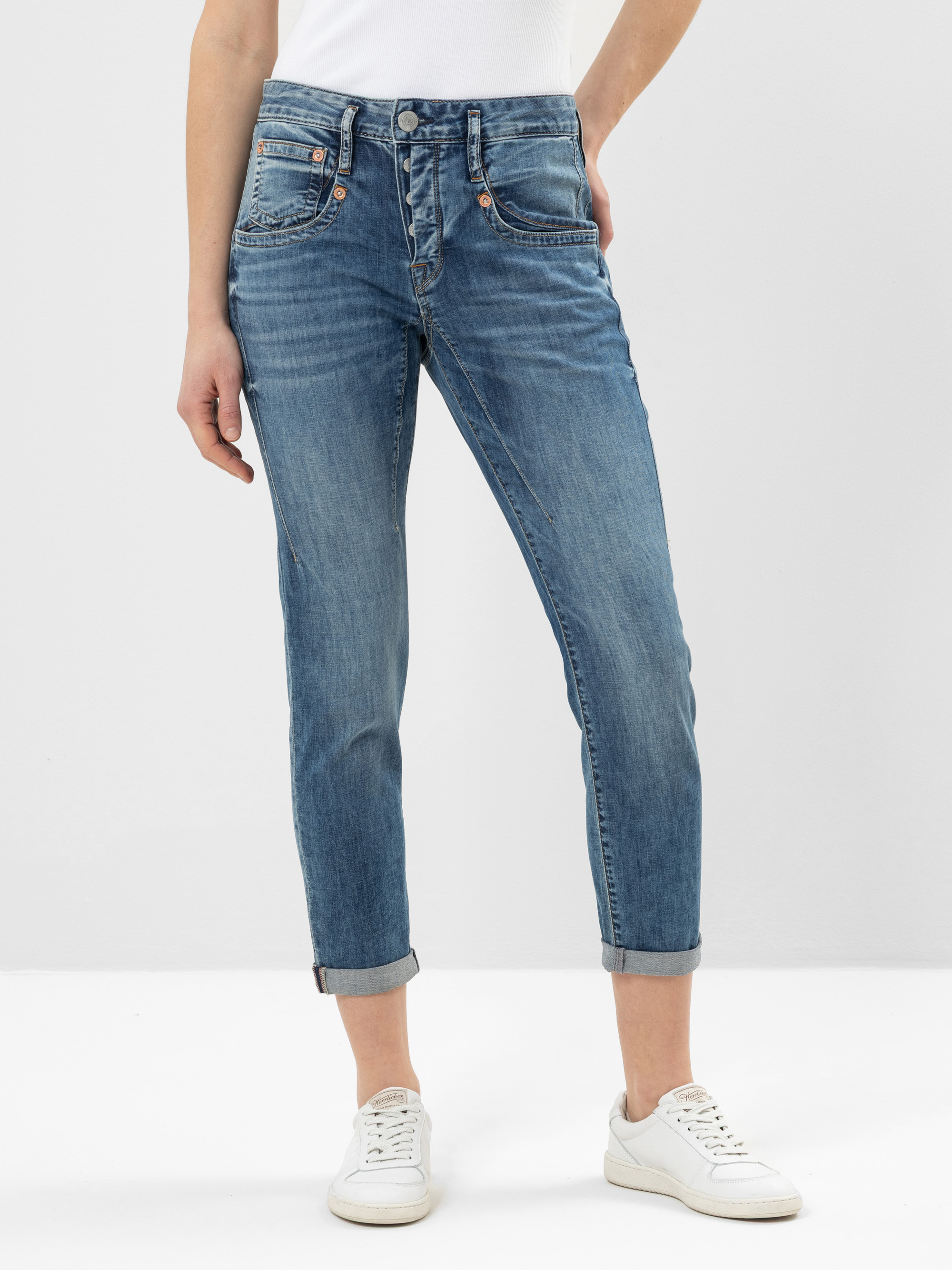 Eine Person, die hochgekrempelte Jeans, ein weißes Oberteil und weiße Turnschuhe trägt, steht vor einem schlichten hellen Hintergrund. Nur der Unterkörper ist sichtbar.
