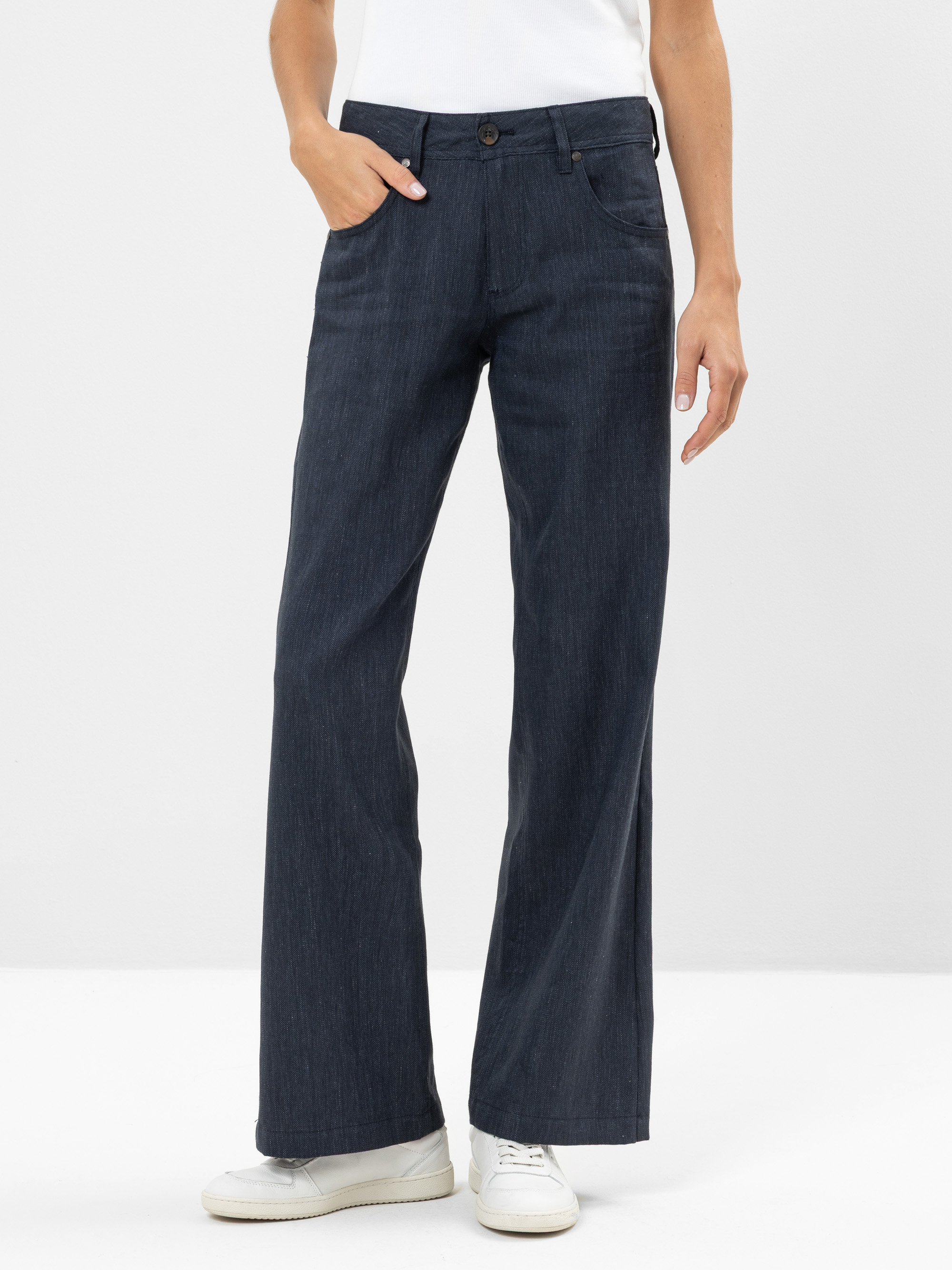 Eine Person, die dunkelblaue Jeans mit weitem Bein, ein weißes Hemd und weiße Turnschuhe trägt, steht mit einer Hand in der Tasche vor einem schlichten hellen Hintergrund. Der obere Teil des Körpers ist aus dem Bild herausgeschnitten.