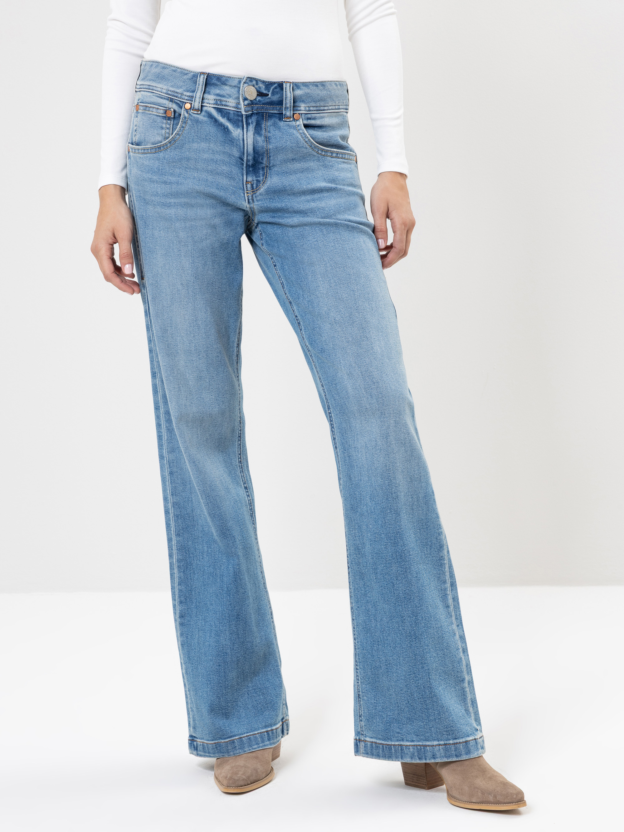 Eine Person, die hellblaue, weit geschnittene Jeans, ein weißes, langärmeliges Oberteil und beigefarbene, hochhackige Stiefel trägt, steht vor einem schlichten, hellen Hintergrund.