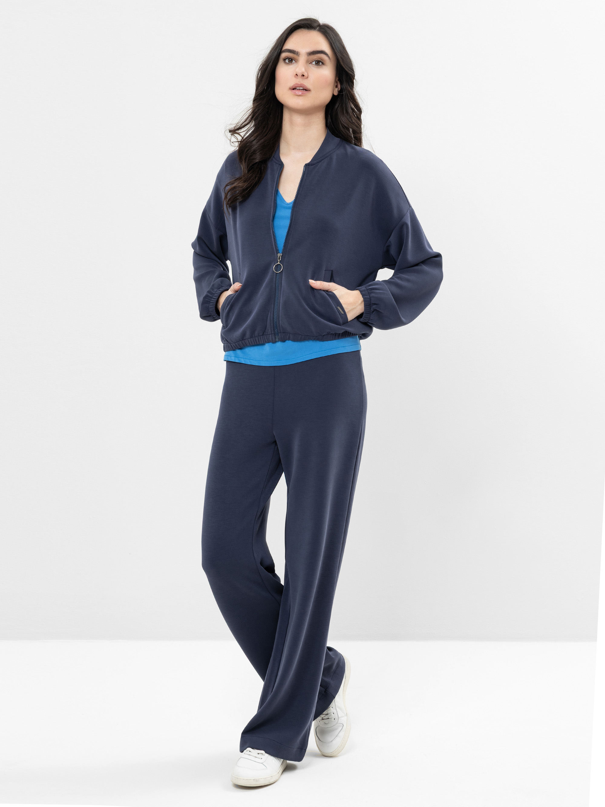 Eine Frau steht vor einem weißen Hintergrund und trägt eine marineblaue Jacke mit Reißverschluss und eine passende Hose mit weitem Bein, darunter ein hellblaues Hemd. Sie hat langes dunkles Haar, Hände in den Jackentaschen und trägt weiße Turnschuhe