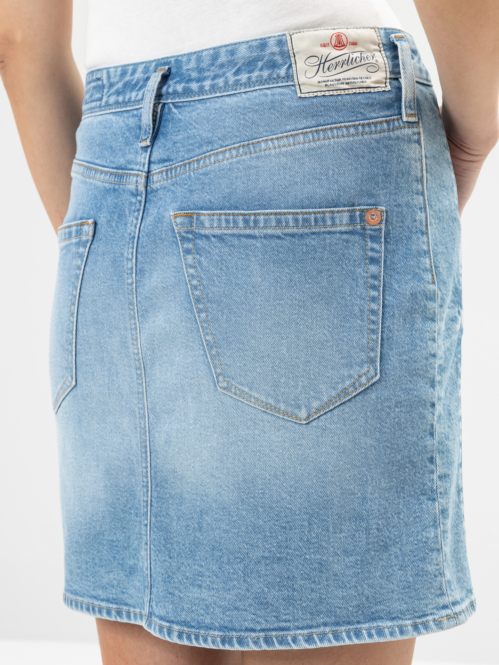 Nahaufnahme einer Person, die einen hellblauen Jeansrock mit Gesäßtaschen und einem weißen Etikett mit der Aufschrift Herrlicher am Bund trägt. Die Person trägt außerdem ein weißes Oberteil.