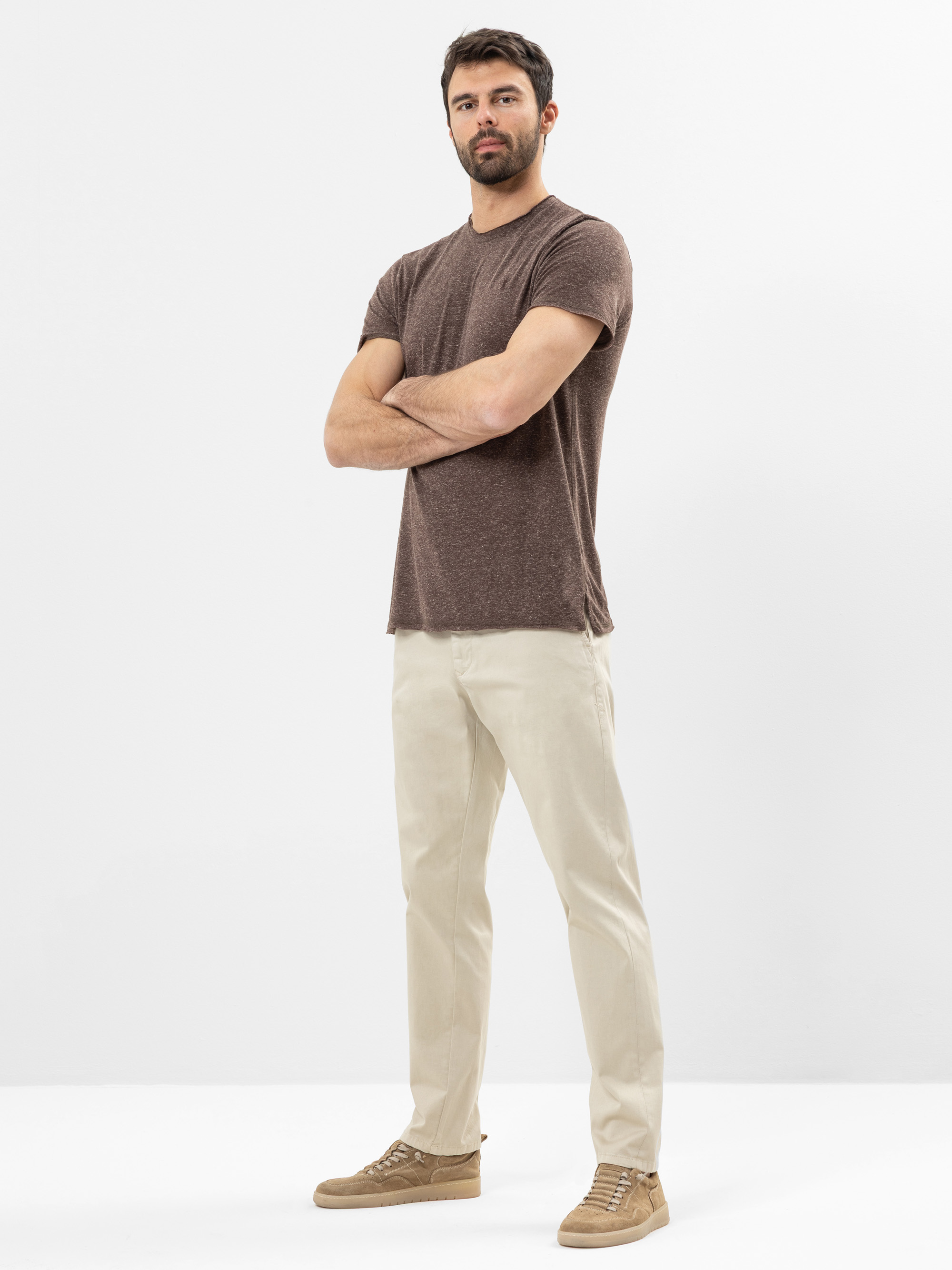 Ein Mann mit kurzen dunklen Haaren und einem Bart steht mit verschränkten Armen, einem braunen T-Shirt, einer beigen Hose und hellbraunen Turnschuhen vor einem schlichten weißen Hintergrund.