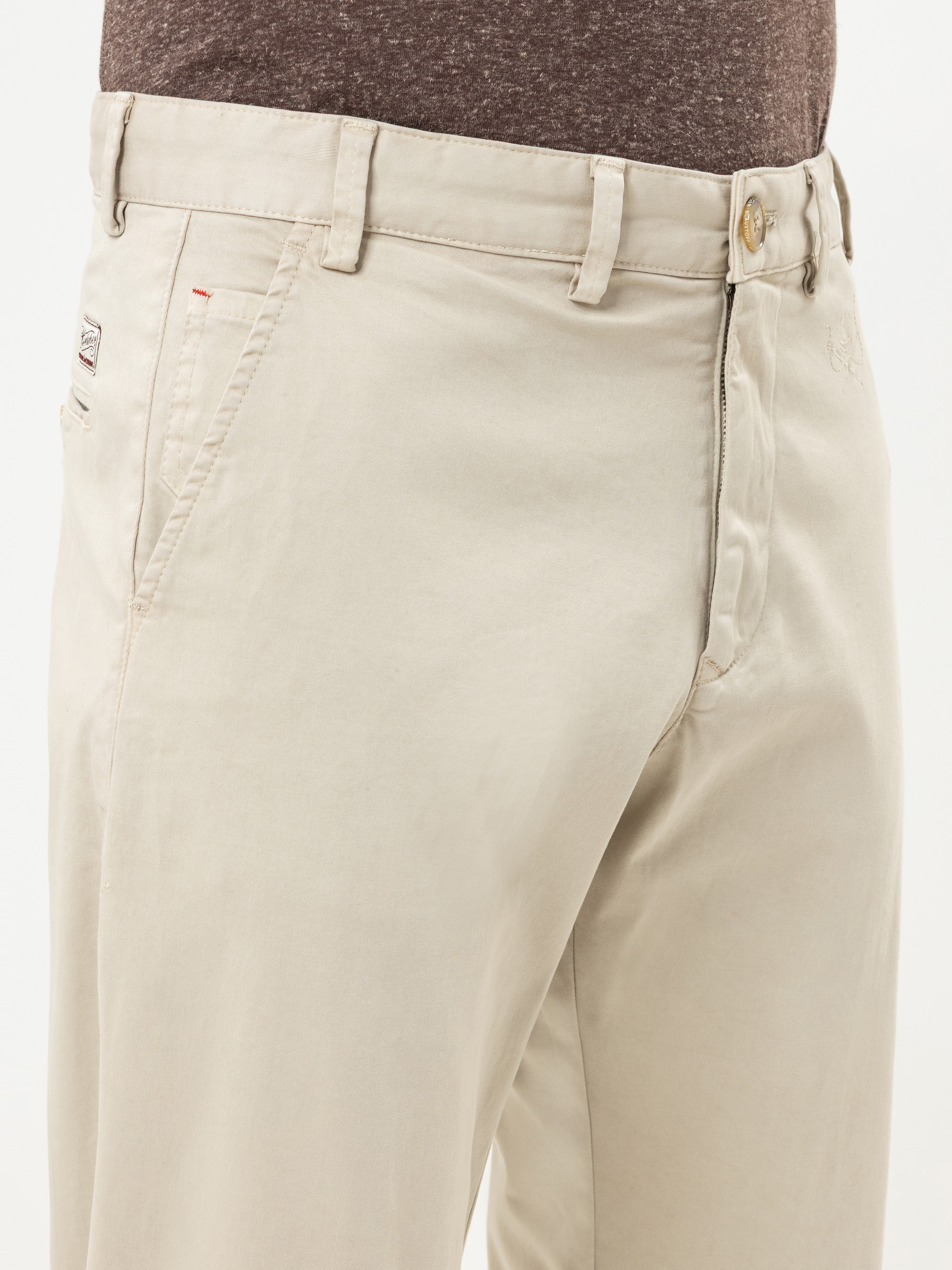 Eine Person, die eine hellbeige Hose mit einem braunen Gürtel und ein braunes Hemd trägt, von der Taille bis zur Mitte des Oberschenkels abgebildet, steht vor einem schlichten Hintergrund.