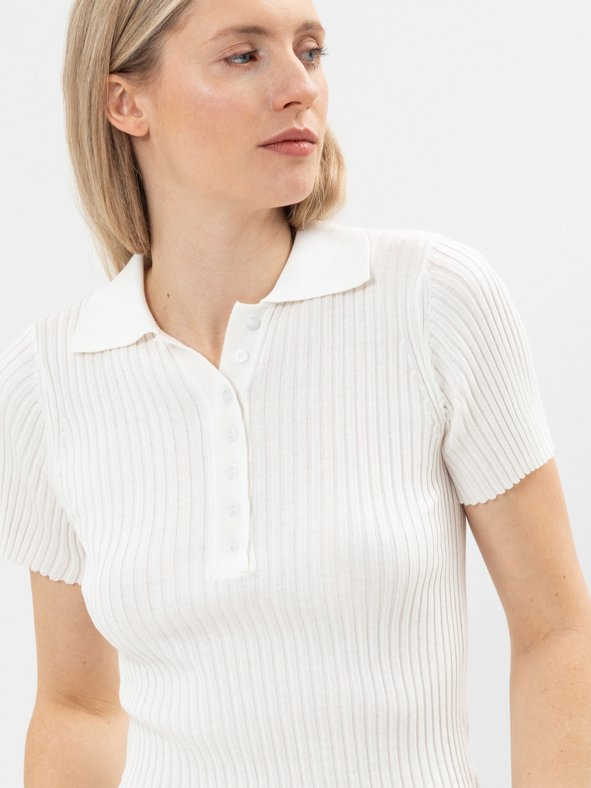 Eine Frau mit blondem Haar, die ein kurzärmeliges, geripptes weißes Poloshirt trägt, blickt vor einem schlichten weißen Hintergrund zur Seite.