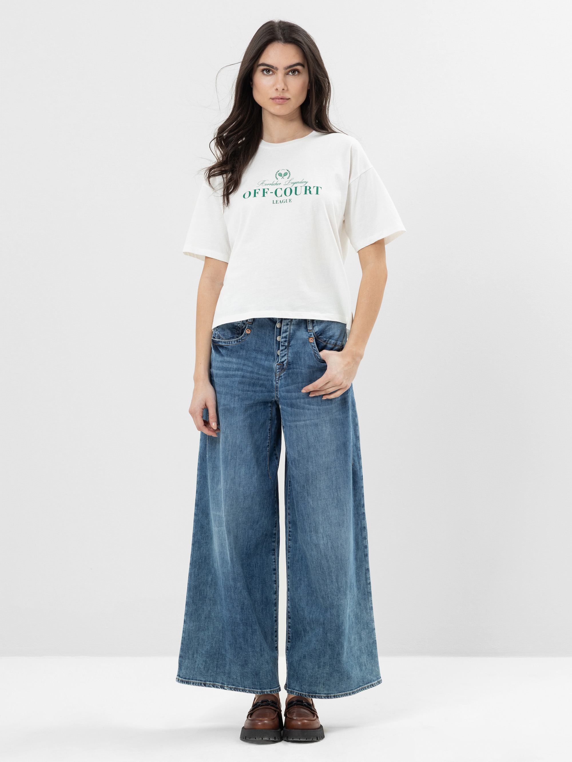 Eine Frau steht vor einem schlichten weißen Hintergrund und trägt ein weißes T-Shirt mit der Aufschrift OFF-COURT, weite Bluejeans und braune Schuhe. Ihr langes dunkles Haar ist offen, und ihre Arme sind locker an den Seiten abgelegt.