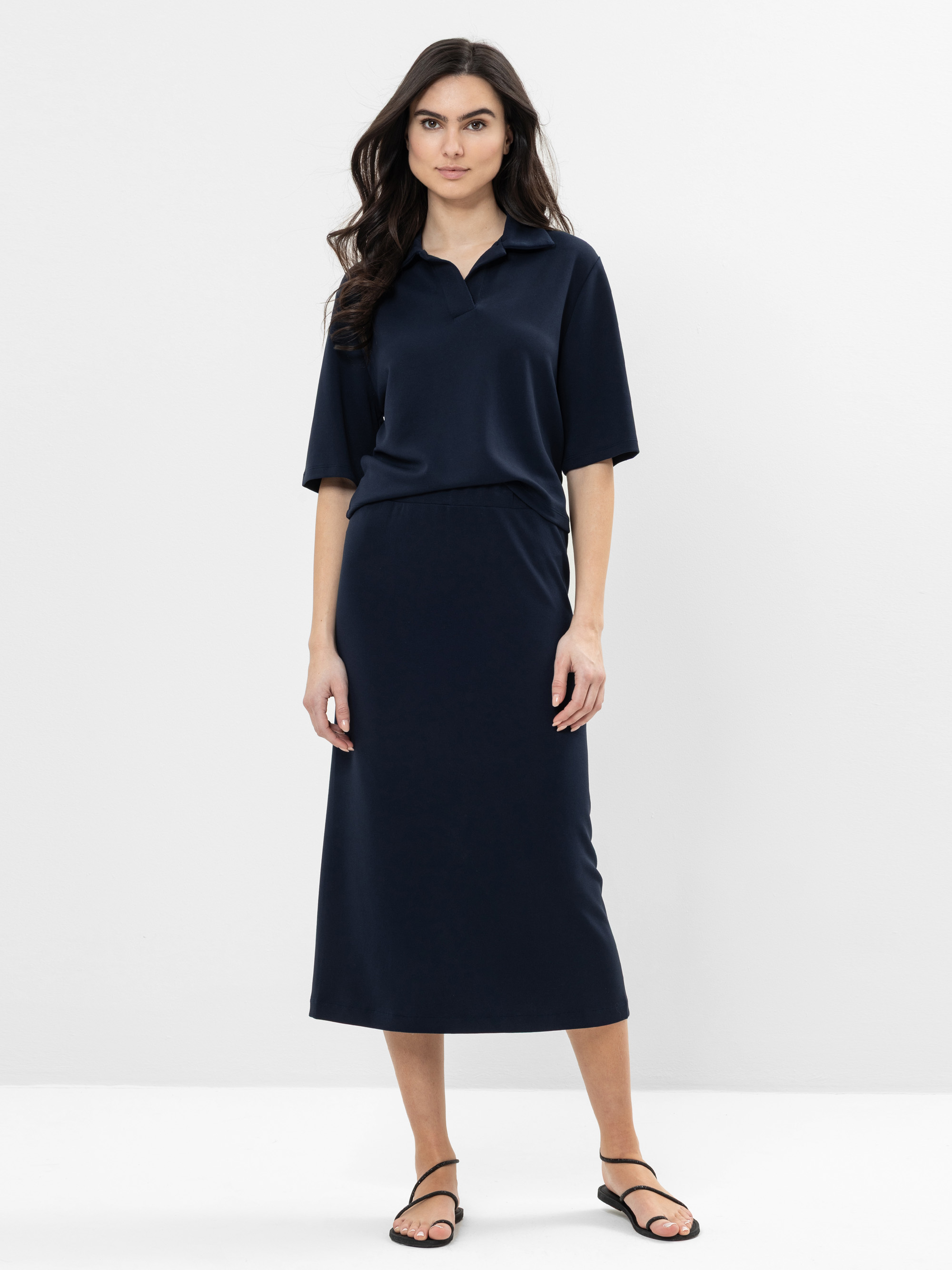 Eine Frau mit langen dunklen Haaren trägt ein marineblaues, kurzärmeliges Midikleid mit Kragen und schwarze Sandalen. Sie steht mit dem Gesicht nach vorne vor einem schlichten weißen Hintergrund.