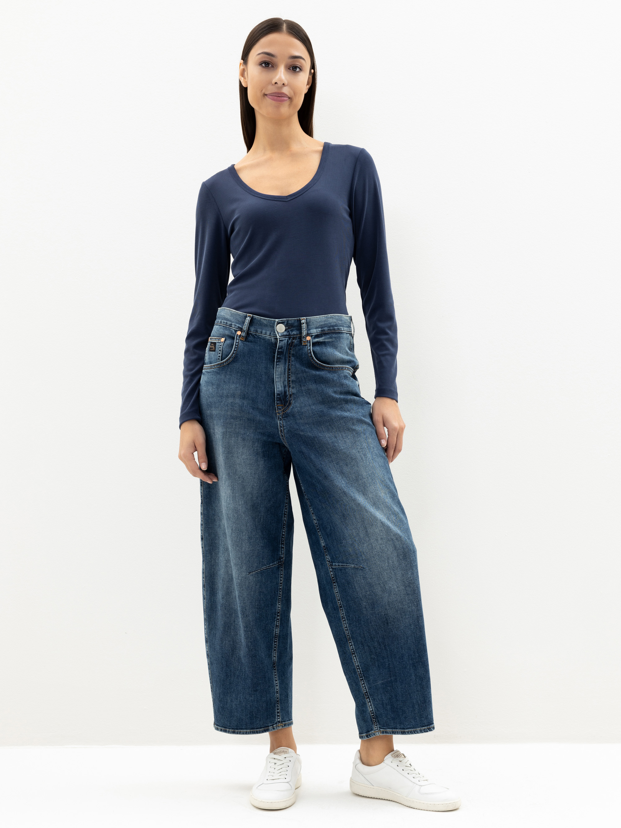 Eine Frau steht vor einem schlichten weißen Hintergrund und trägt ein marineblaues langärmeliges Oberteil mit Rundhalsausschnitt, weite Bluejeans und weiße Turnschuhe. Sie hat glattes, dunkles Haar und eine entspannte, selbstbewusste Körperhaltung.