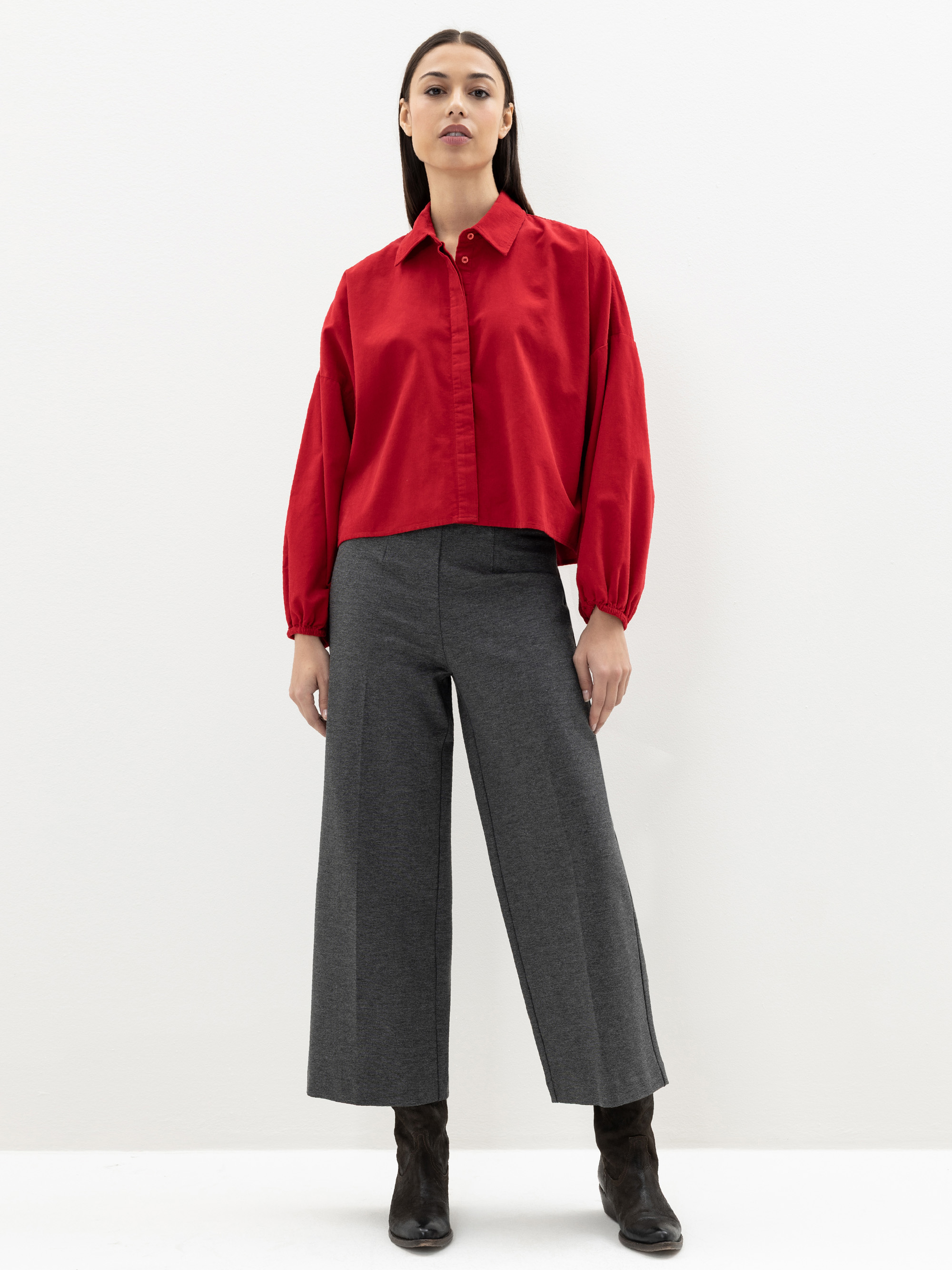 Eine Frau steht vor einem schlichten weißen Hintergrund und trägt ein locker sitzendes rotes Button-up-Hemd, eine graue Hose mit weitem Bein und schwarze Stiefel. Sie blickt nach vorne und hat die Arme entspannt an den Seiten.