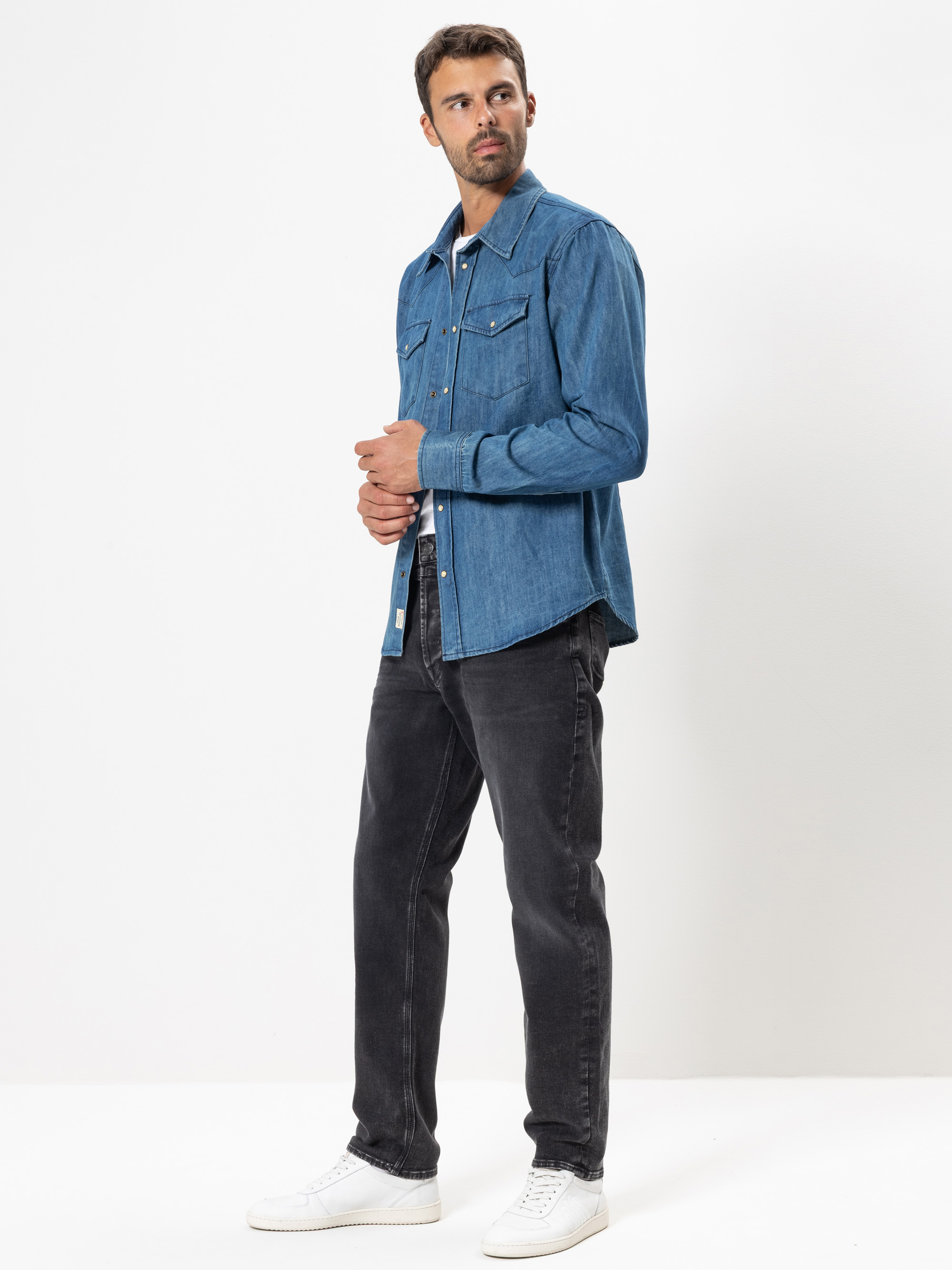 Ein Mann steht vor einem schlichten weißen Hintergrund und trägt ein blaues Jeanshemd, schwarze Jeans und weiße Turnschuhe. Er schaut leicht zur Seite und hat die Hände in der Nähe der Hemdknöpfe.