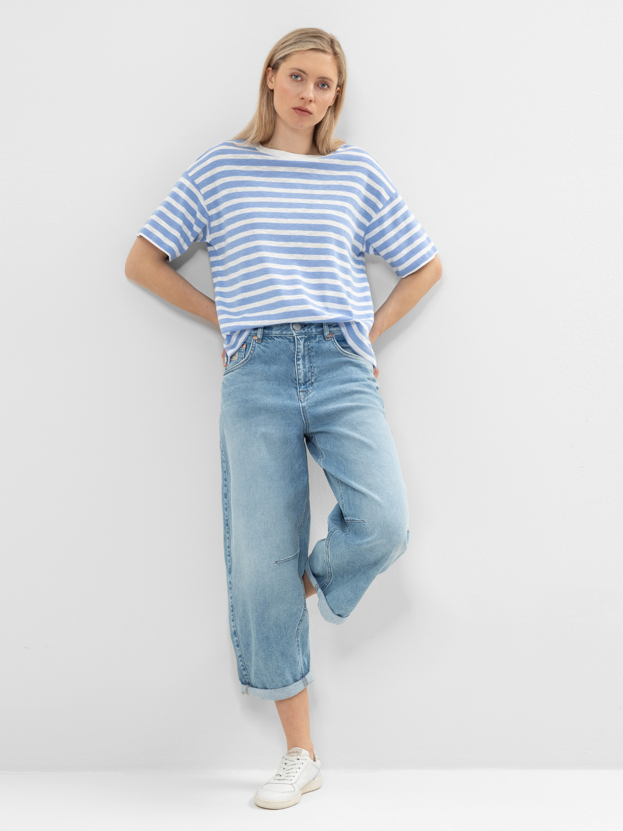 Eine Frau steht vor einer weißen Wand und trägt ein hellblau-weiß gestreiftes T-Shirt, hellblaue Jeans mit Bündchen und weiße Turnschuhe. Sie hat glattes blondes Haar und ihre Hände stecken in den Taschen ihrer Jeans.