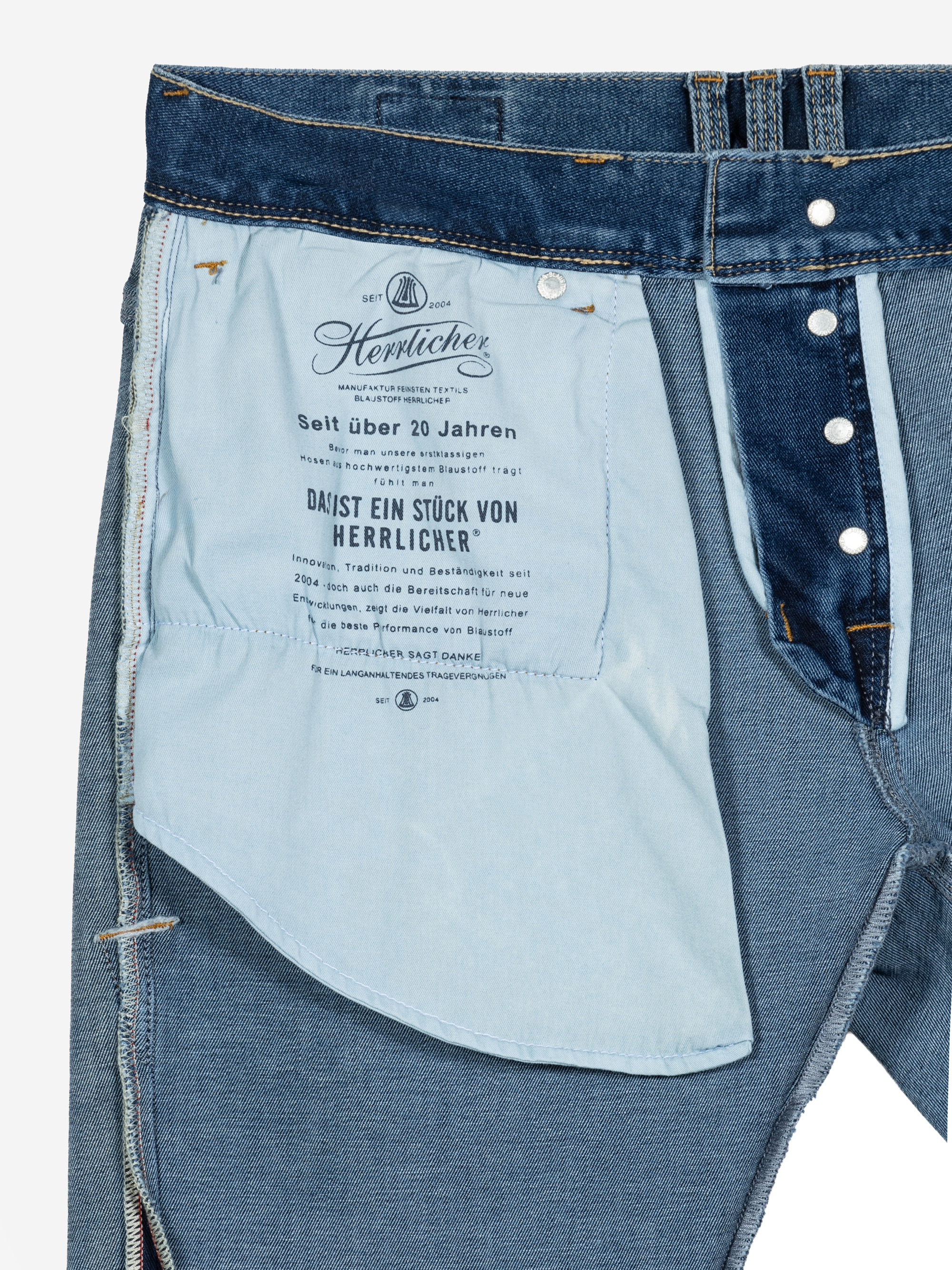 Eine Nahaufnahme der Innenseite einer blauen Jeans, die ein großes hellblaues Taschenfutter mit aufgedrucktem deutschen Text zeigt. Die Jeans ist teilweise aufgeknöpft, um die inneren Details und Nähte zu zeigen.