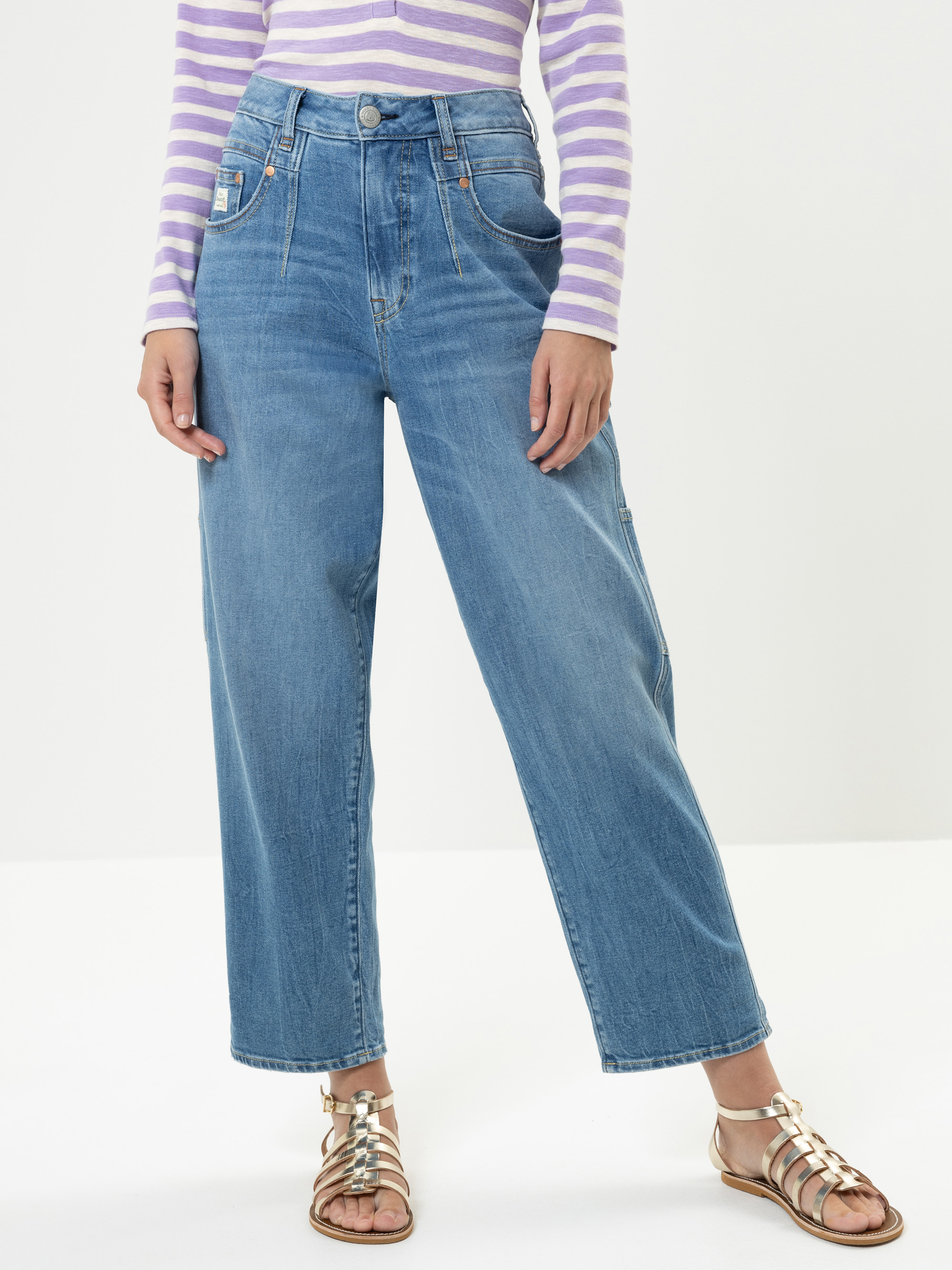 Eine Person, die hoch taillierte, weit geschnittene Bluejeans, ein langärmeliges, lavendelfarben gestreiftes Oberteil und metallische Riemchensandalen trägt, steht vor einem schlichten hellen Hintergrund.