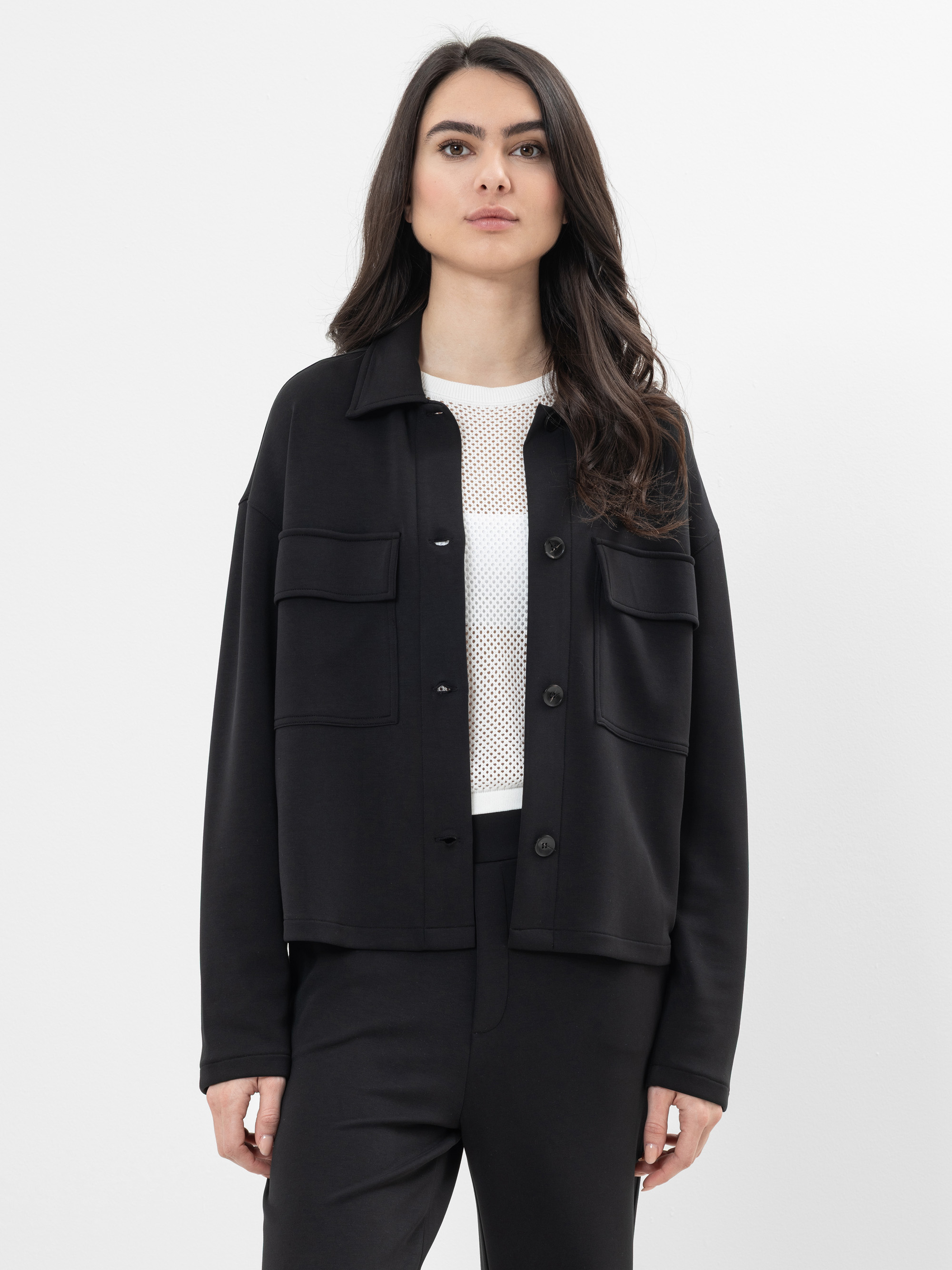 Eine Frau mit langen dunklen Haaren, die eine schwarze Jacke mit Knöpfen und großen Vordertaschen über einem weißen Netzoberteil und einer schwarzen Hose trägt, steht vor einem schlichten weißen Hintergrund und schaut geradeaus.