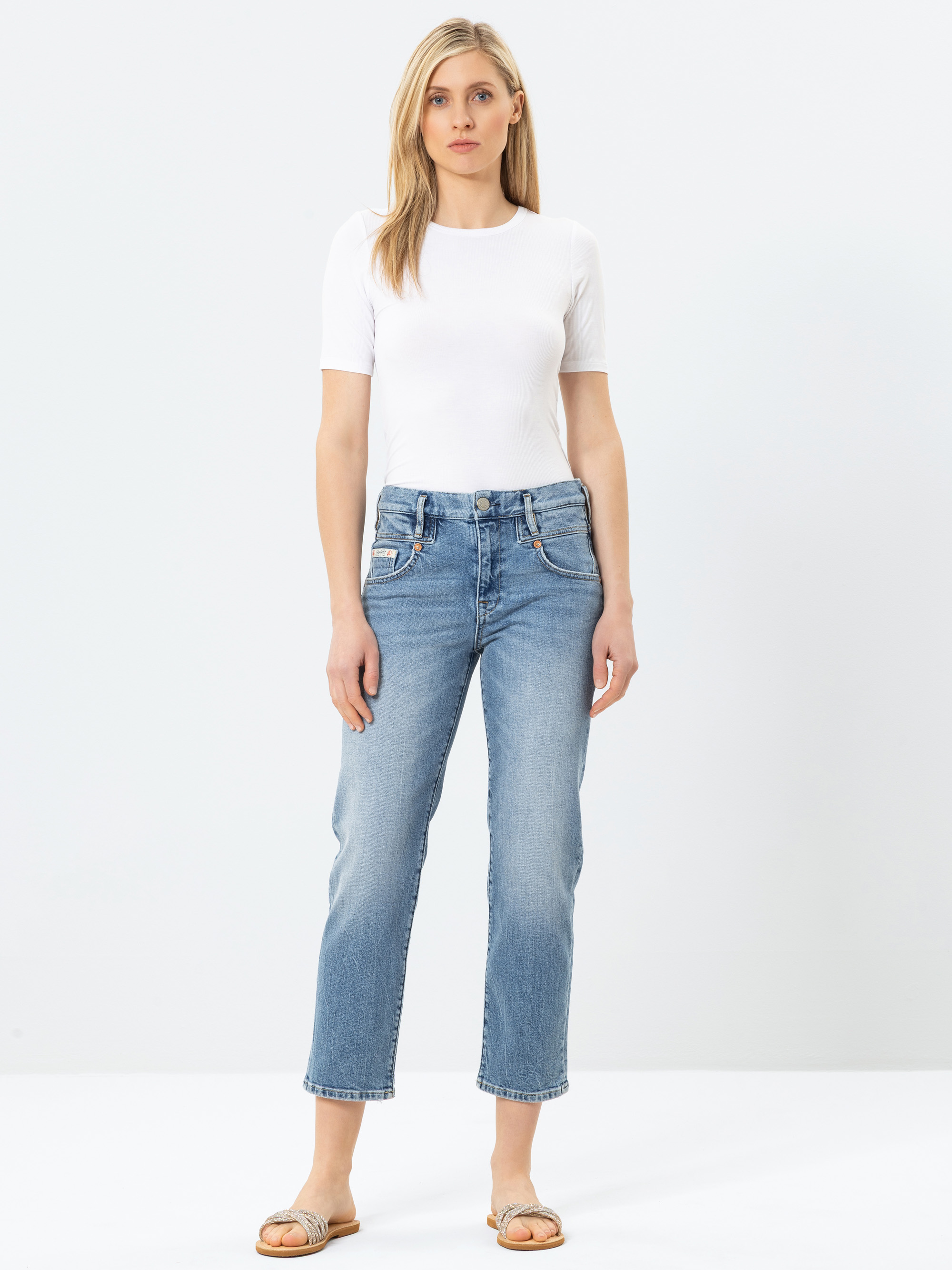 Eine Frau steht mit dem Gesicht nach vorne, trägt ein kurzärmeliges weißes Oberteil, hellblaue Jeans mit hoher Taille und hellbraune Sandalen vor einem schlichten weißen Hintergrund.