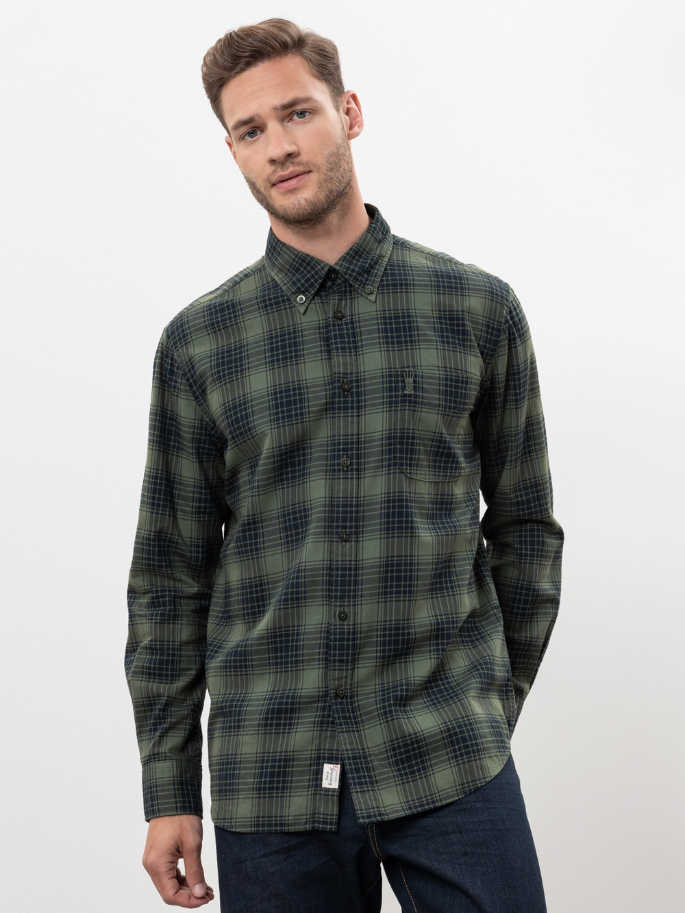 Ein Mann, der ein grün und marineblau kariertes Button-up-Hemd und dunkle Jeans trägt, steht vor einem schlichten weißen Hintergrund und blickt mit einem neutralen Ausdruck leicht zur Seite.