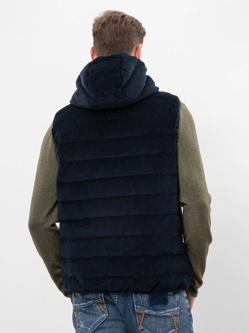 Eine Person, die eine marineblaue Steppweste mit Kapuze, ein olivgrünes langärmeliges Hemd und eine blaue Jeans trägt, wird von hinten vor einem schlichten weißen Hintergrund gezeigt.