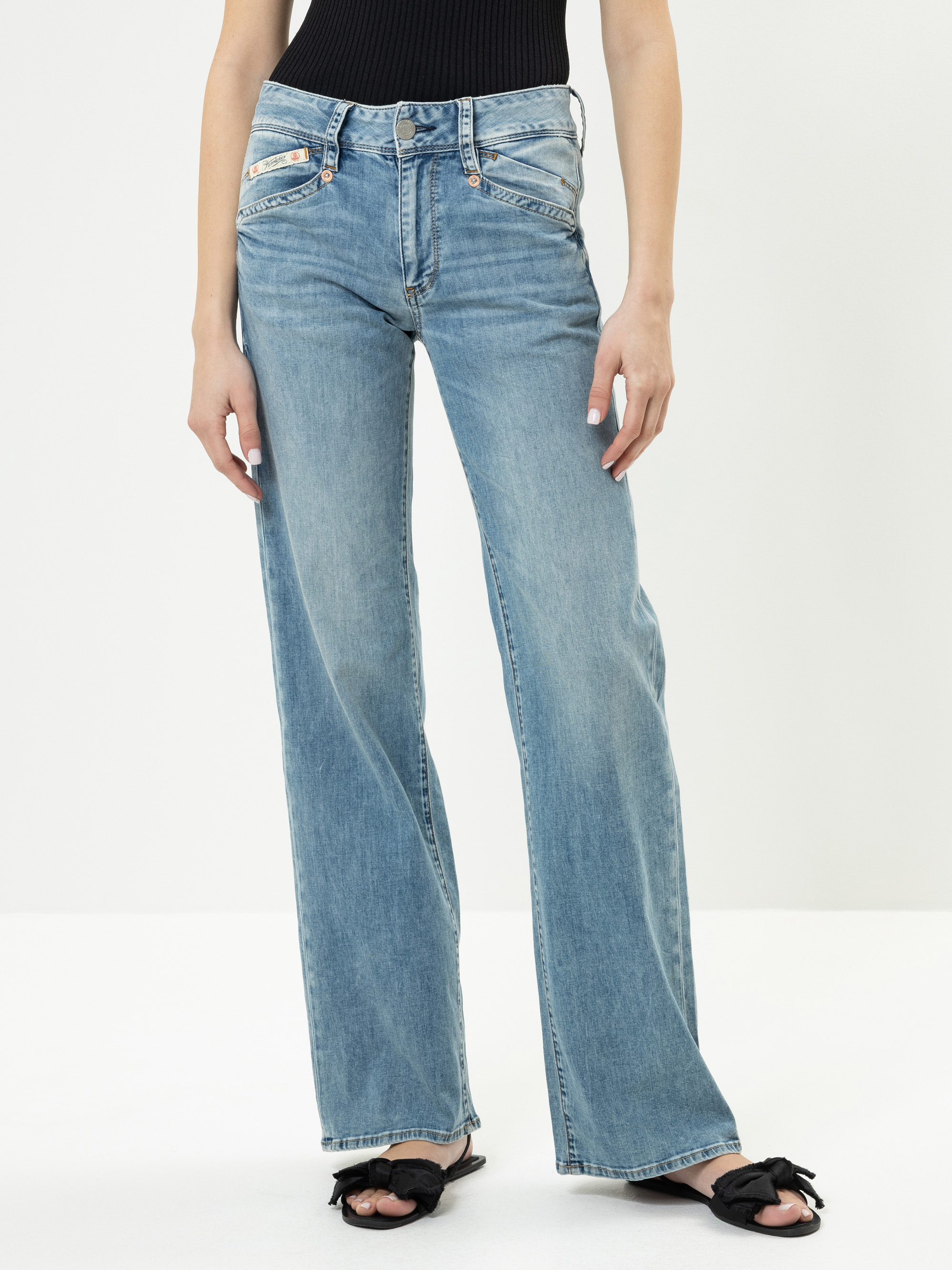 Eine Person, die hellblaue Jeans mit geradem Bein, ein schwarzes geripptes ärmelloses Oberteil und schwarze Gleitsandalen mit Schleifen trägt, steht vor einem schlichten weißen Hintergrund.
