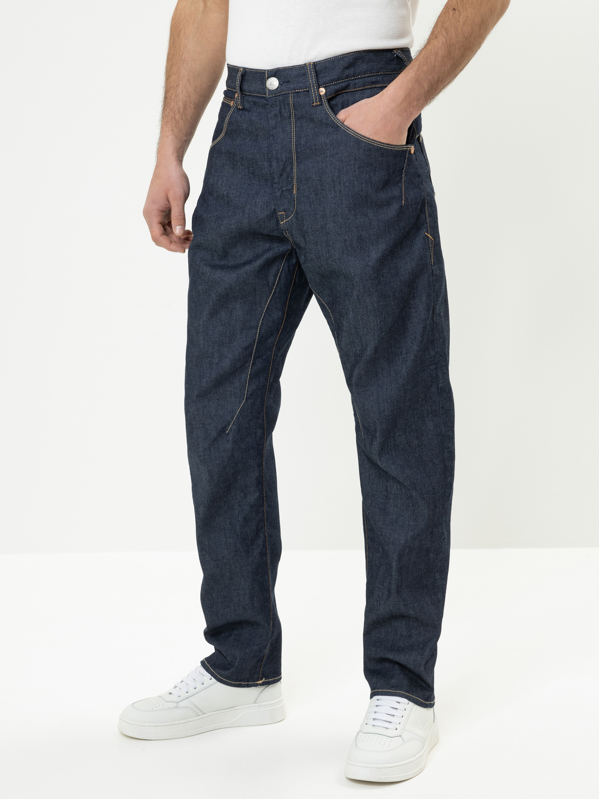 Eine Person, die dunkelblaue Jeans mit geradem Bein und orangefarbenen Nähten, ein weißes T-Shirt und weiße Turnschuhe trägt, steht vor einem schlichten weißen Hintergrund und hat eine Hand in der Tasche.