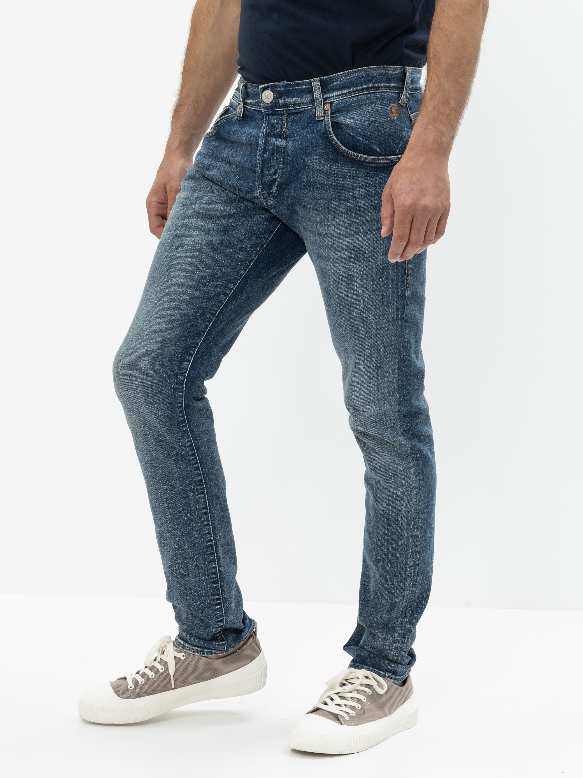 Ein Mann, der ein schwarzes T-Shirt, blaue Slim-Fit-Jeans und hellgraue Turnschuhe mit weißen Sohlen trägt, steht vor einem schlichten weißen Hintergrund. Sein Kopf ist auf dem Bild nicht zu sehen.