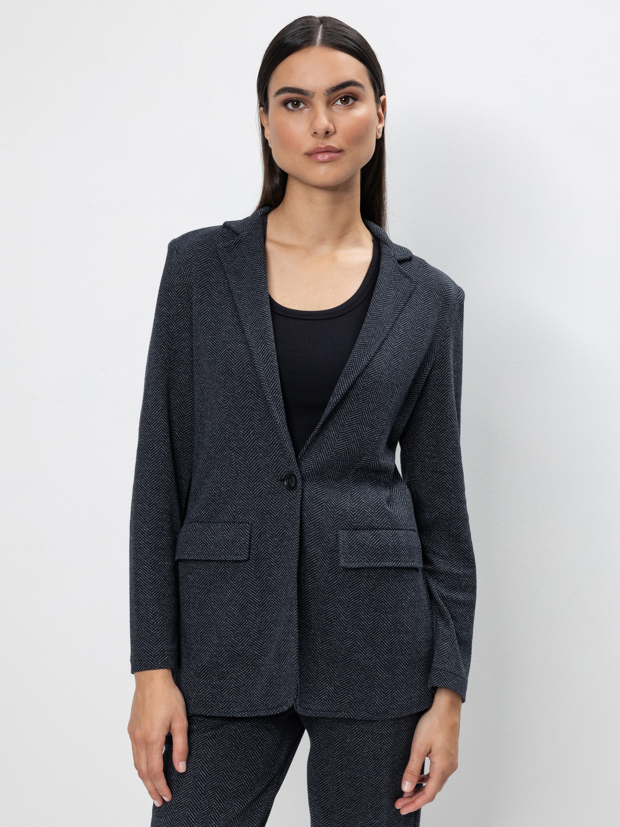 Eine Frau mit glattem dunklem Haar trägt einen dunkelgrauen Blazer über einem schwarzen Oberteil, gepaart mit einer passenden Hose, und steht vor einem schlichten weißen Hintergrund. Sie blickt selbstbewusst und mit neutraler Miene in die Kamera.