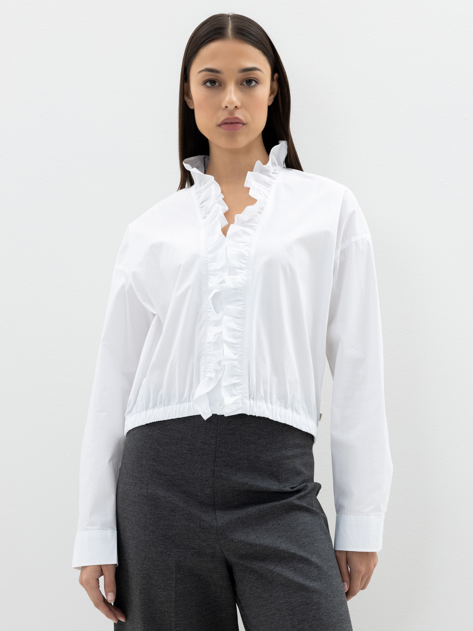Eine Frau steht vor einem schlichten weißen Hintergrund. Sie trägt eine weiße Bluse mit Rüschenbesatz auf der Vorderseite und am Kragen, dazu eine dunkelgraue Hose. Sie hat langes, glattes dunkles Haar und einen neutralen Gesichtsausdruck.