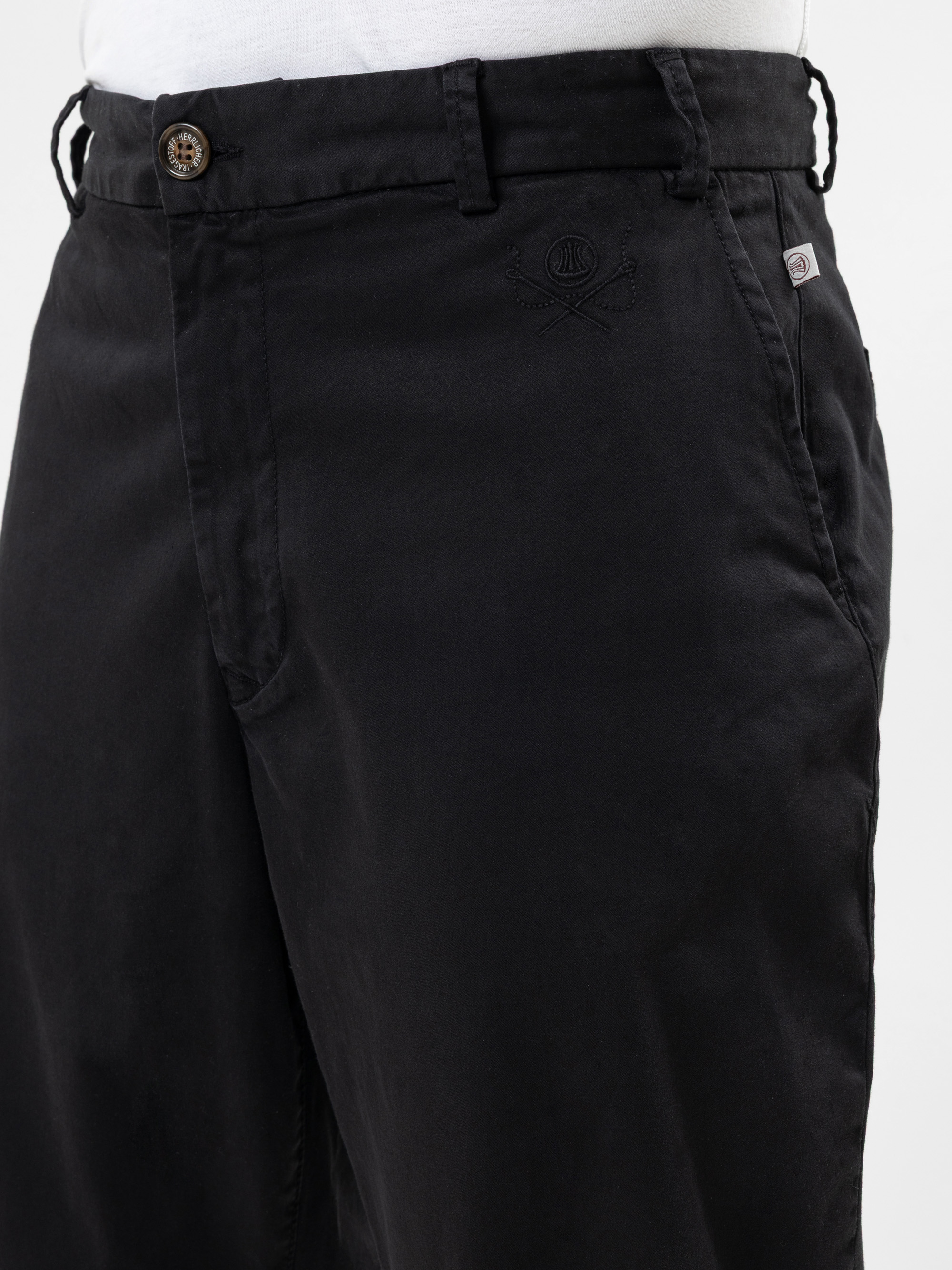 Nahaufnahme einer Person, die eine schwarze Hose mit Knopfverschluss und dezentem gesticktem Logo in der Nähe der Tasche sowie ein weißes Hemd trägt, das in der Taille gesteckt wird.