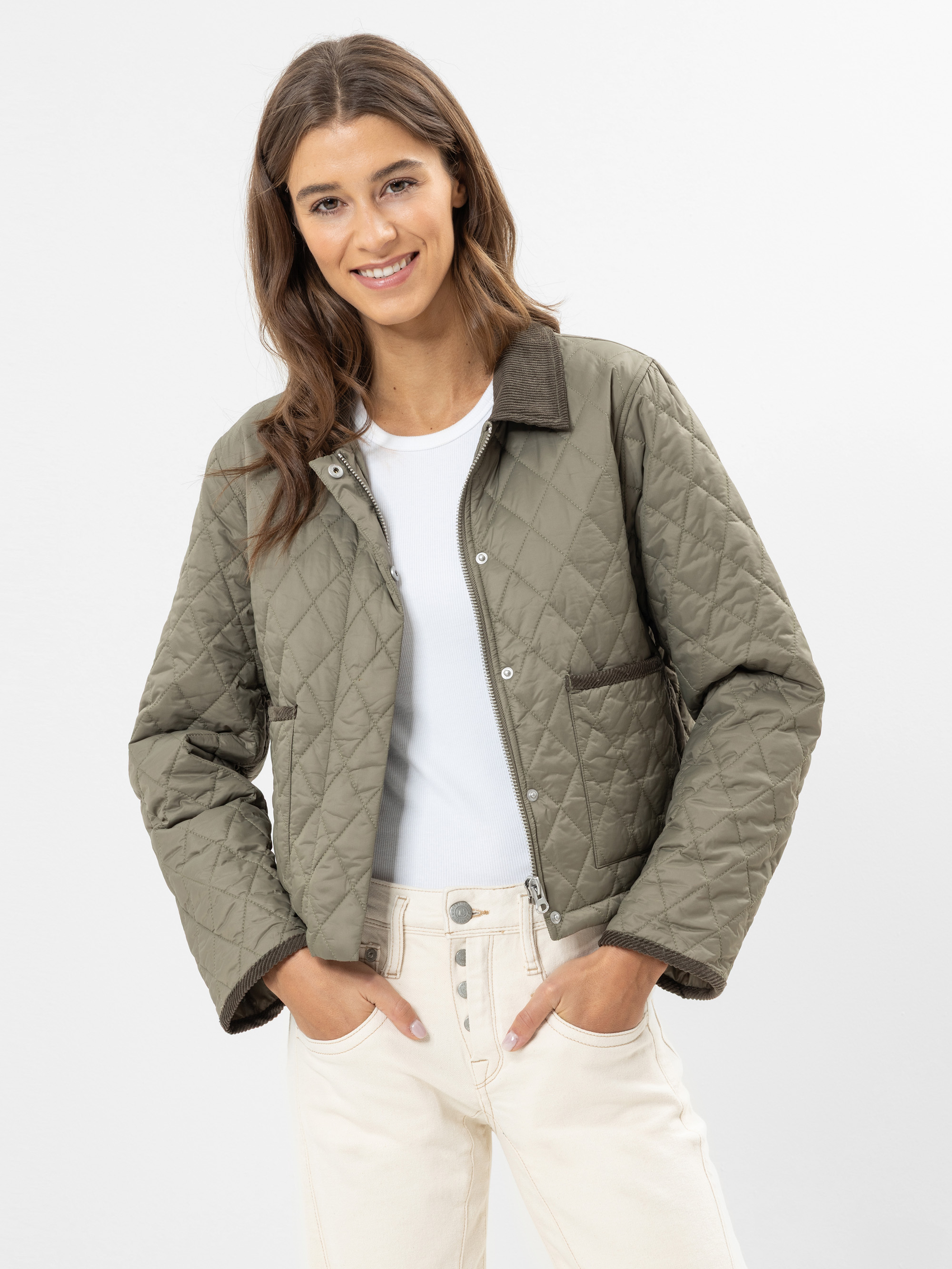 Eine Frau mit langen braunen Haaren lächelt, während sie eine hellgrüne Steppjacke über einem weißen T-Shirt und cremefarbenen Jeans mit hoher Taille trägt. Sie steht vor einem schlichten weißen Hintergrund und hat die Hände in den Taschen.