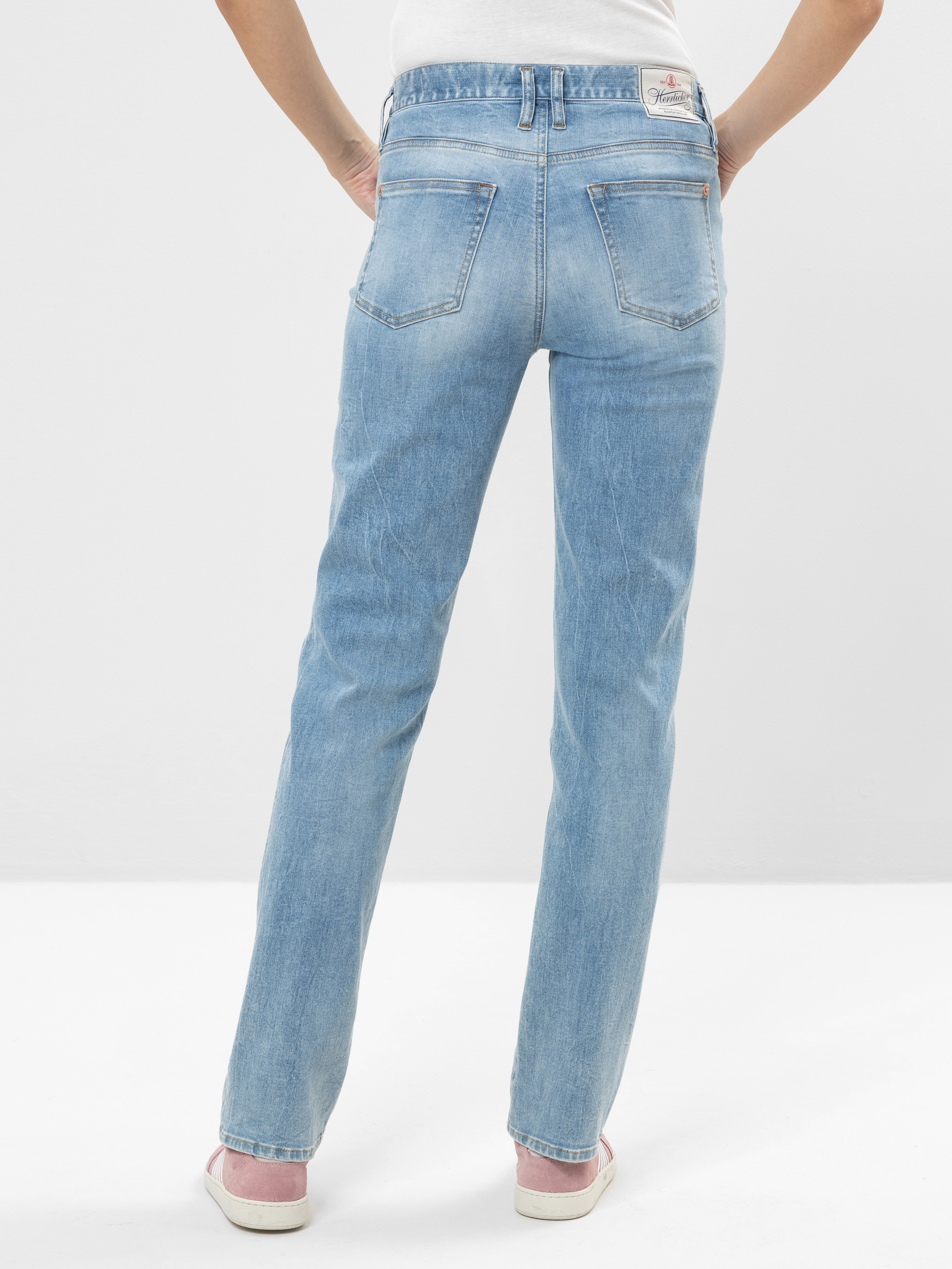 Die Person steht mit dem Rücken zur Kamera und trägt eine hellblaue Jeans mit geradem Bein und rosa Turnschuhe. Die Jeans hat Gesäßtaschen und ein Etikett am Bund. Der Hintergrund ist einfarbig und hell gefärbt.