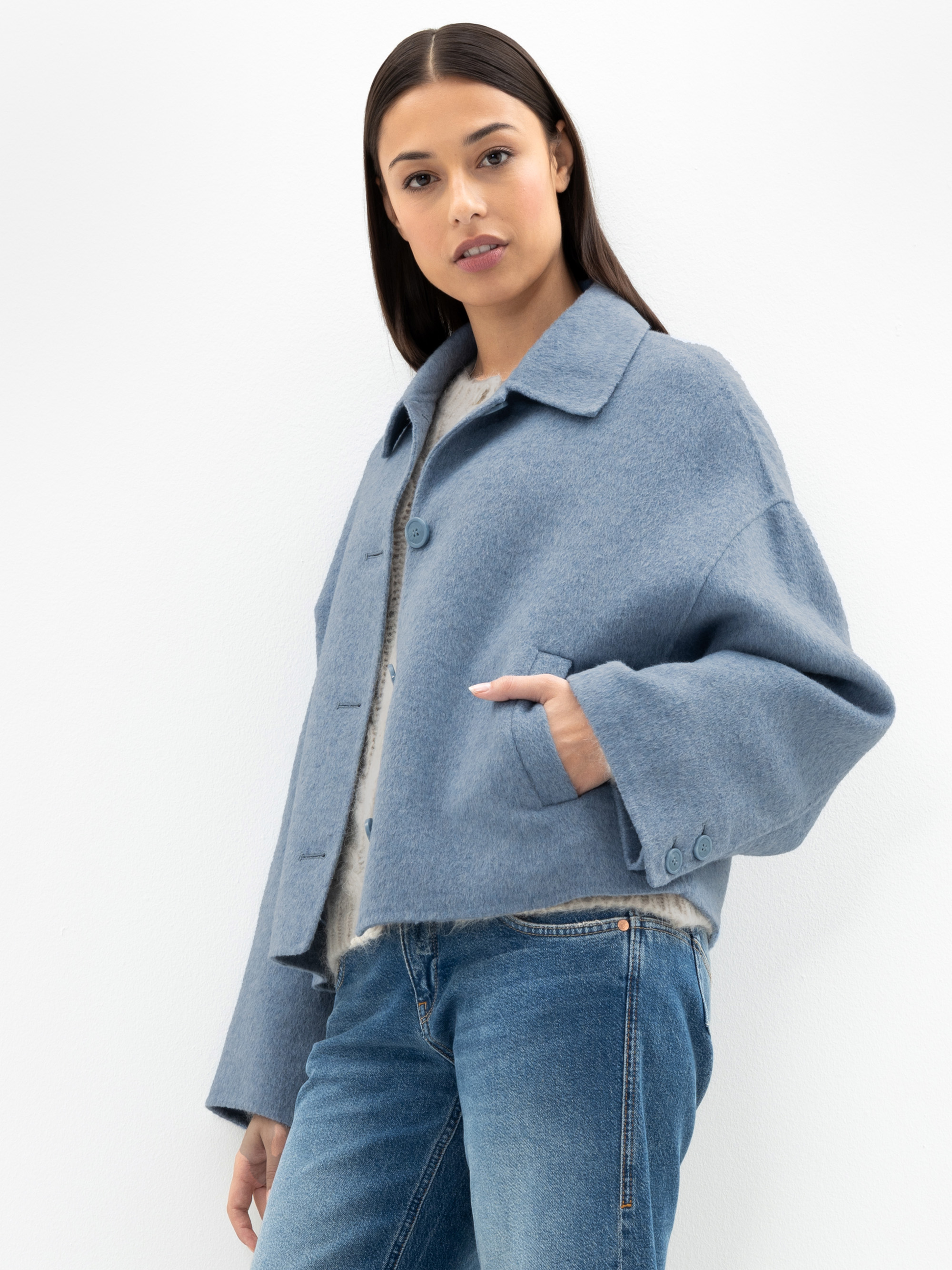 Eine Frau mit glattem dunklem Haar trägt eine hellblaue, geknöpfte Wolljacke und blaue Jeans und steht vor einem schlichten weißen Hintergrund, eine Hand in der Tasche.
