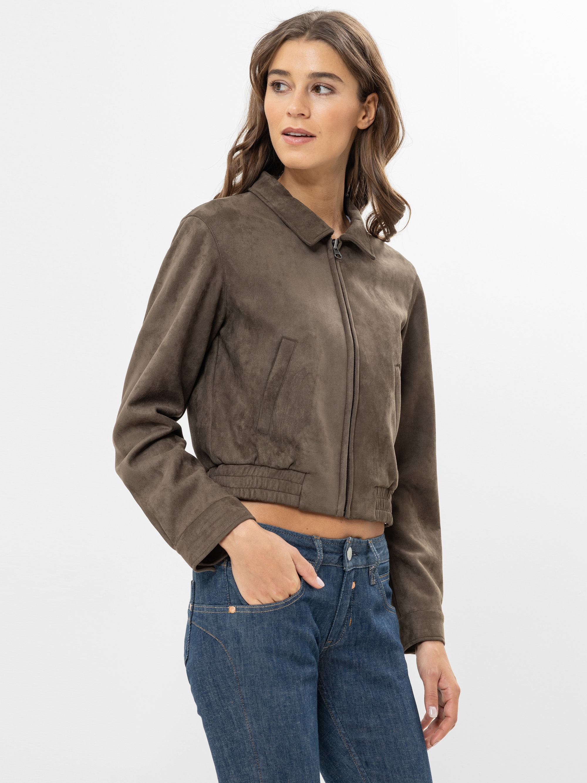 Eine Frau, die eine braune, kurz geschnittene Jacke mit Reißverschluss und eine blaue Jeans trägt, steht vor einem schlichten, hellen Hintergrund und blickt mit einer Hand in ihrer Tasche zur Seite.