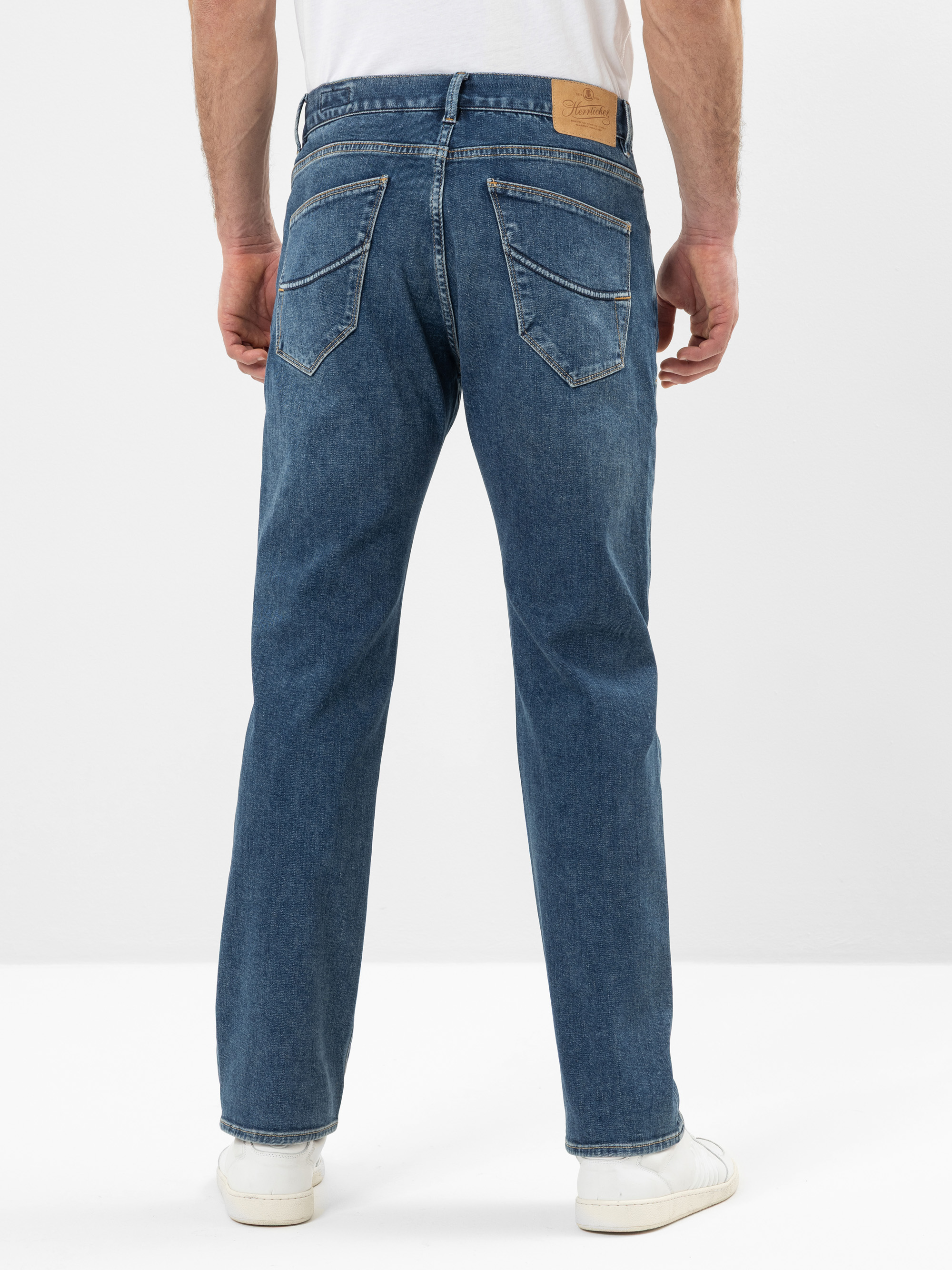 Eine Person, die eine blaue Jeans mit geradem Bein, ein weißes T-Shirt und weiße Turnschuhe trägt, steht mit dem Gesicht nach hinten und zeigt die Rückansicht der Jeans mit zwei Gesäßtaschen und einem hellbraunen Lederaufnäher am Bund.