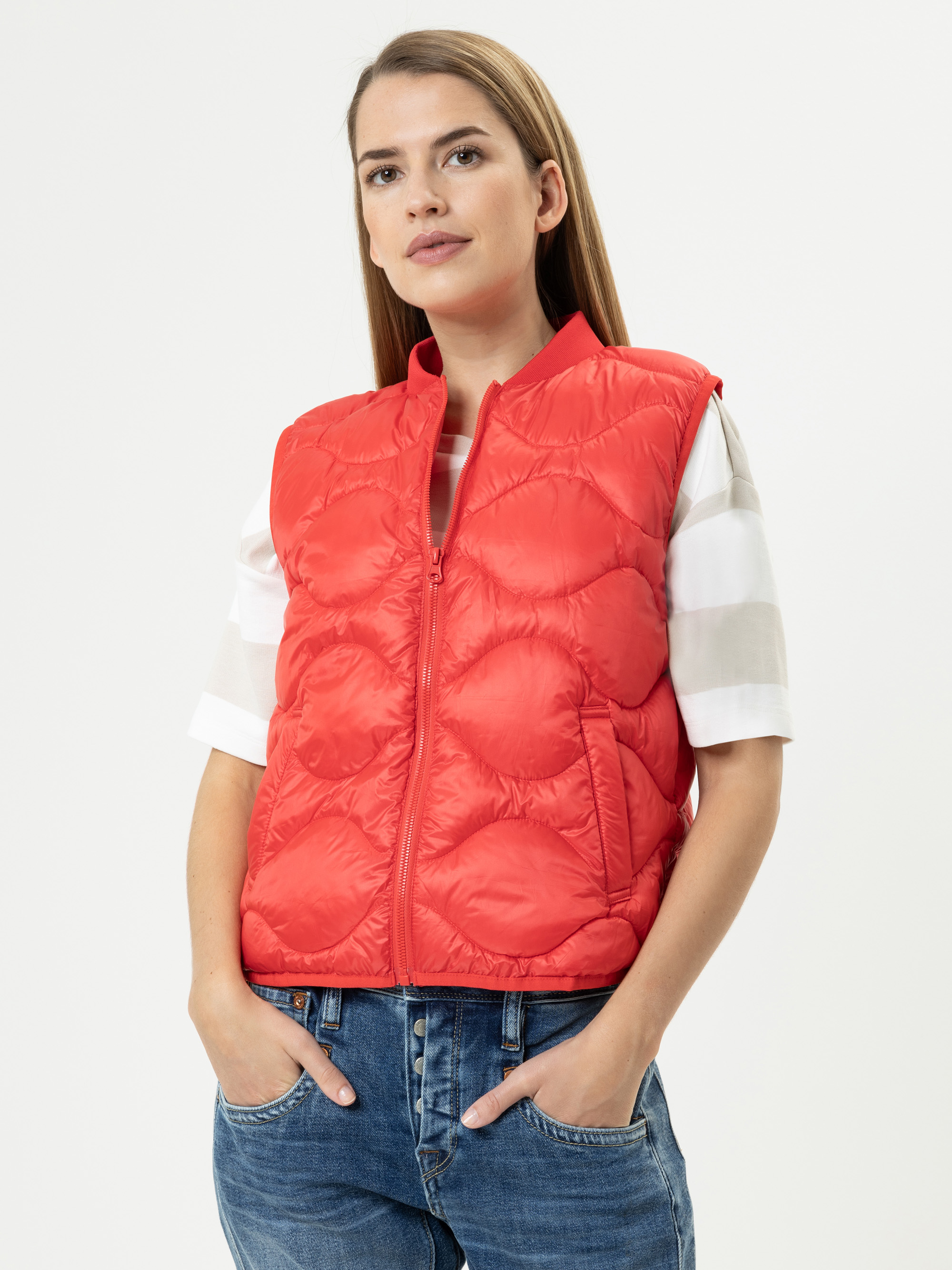 Eine Frau, die eine leuchtend rote ärmellose Steppweste über einem weißen kurzärmeligen Hemd mit breiten Streifen und blauen Jeans trägt, posiert mit den Händen in den Taschen vor einem schlichten weißen Hintergrund.