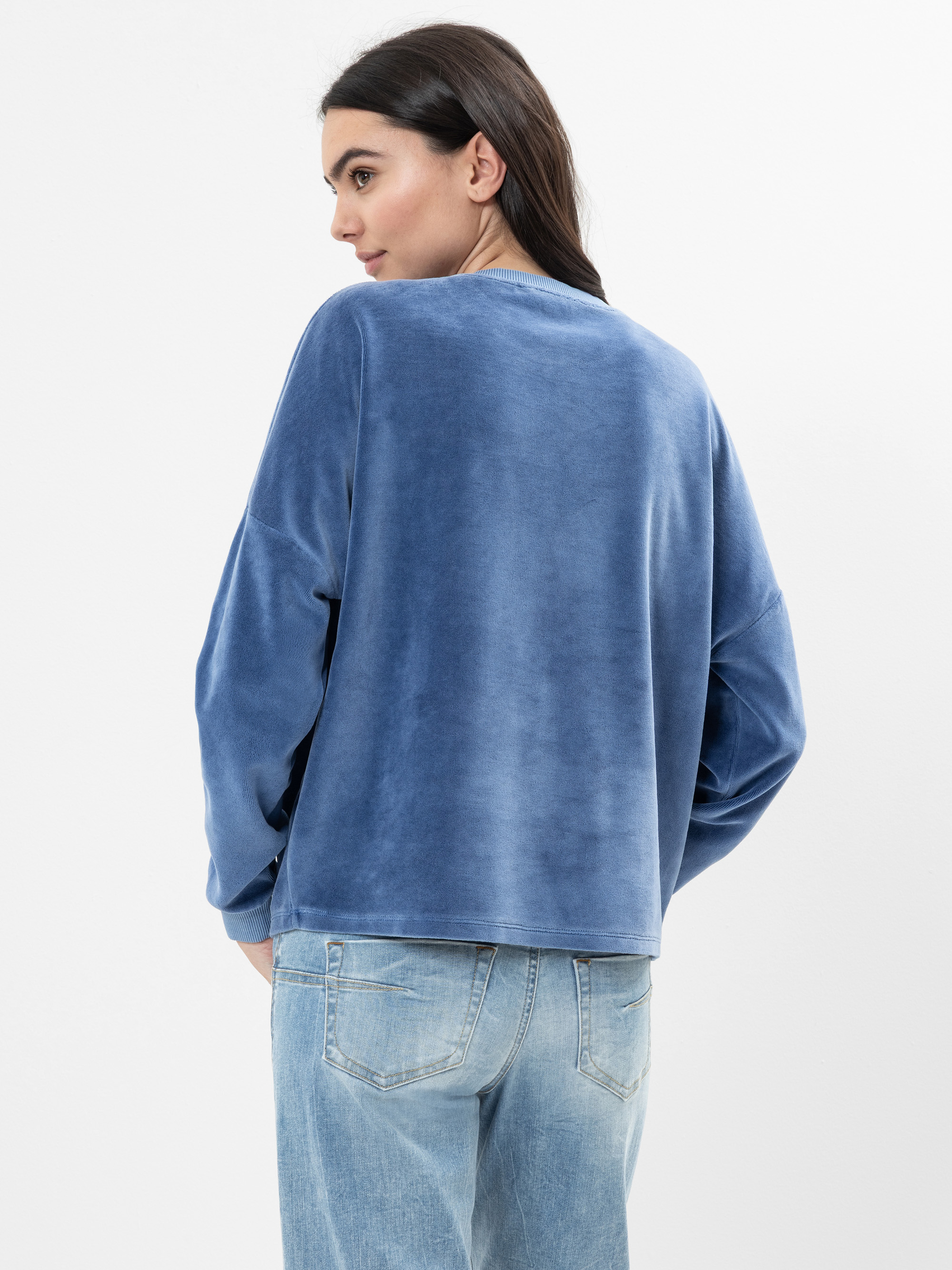Eine Frau mit langen dunklen Haaren steht seitlich und schaut über ihre Schulter. Sie trägt ein zartblaues langärmeliges Oberteil und hellblaue Jeans vor einem schlichten weißen Hintergrund.