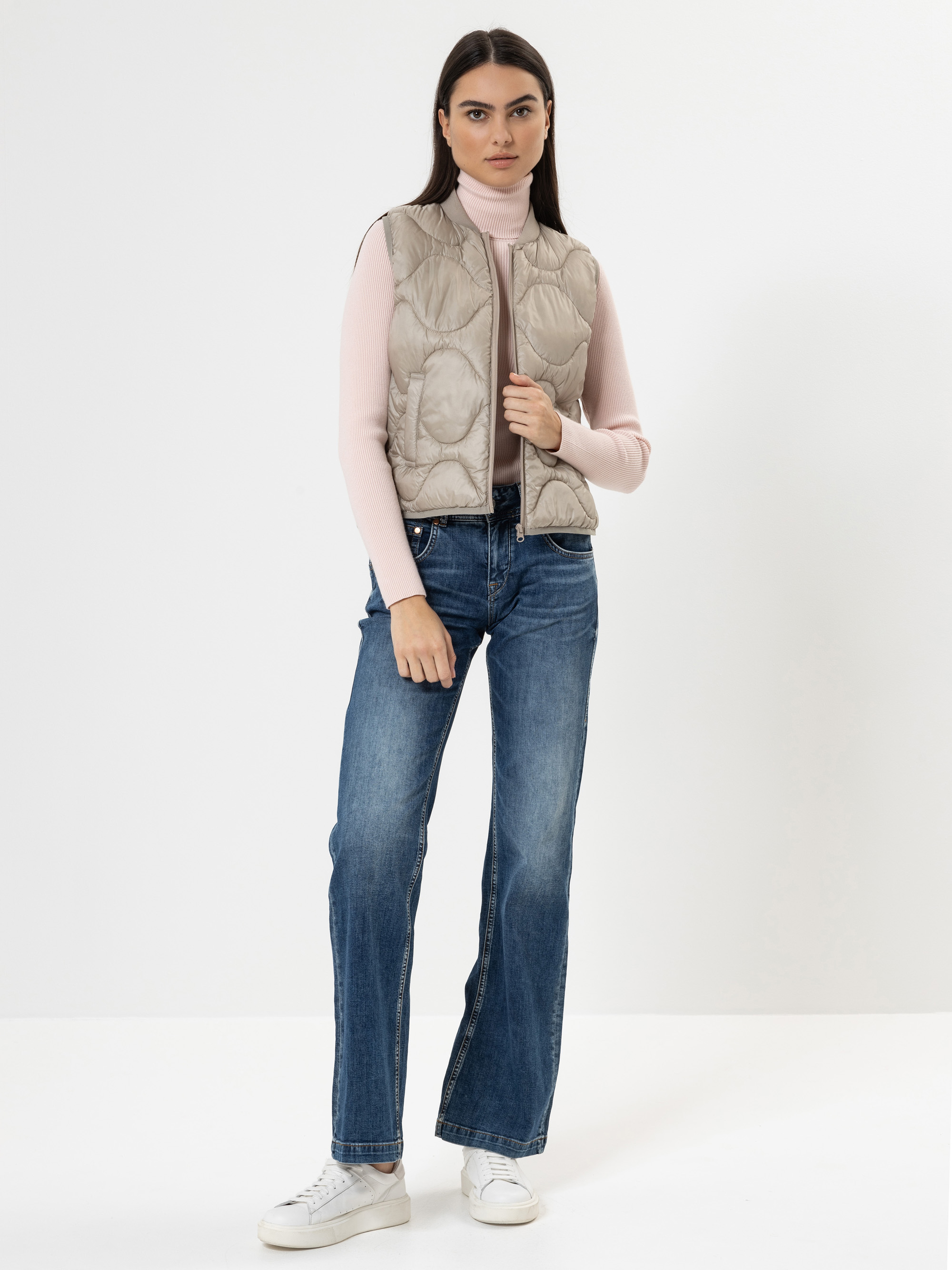 Eine Frau mit langen dunklen Haaren trägt einen hellrosa Rollkragenpullover, eine beige Steppweste, blaue Jeans und weiße Turnschuhe und steht vor einem schlichten weißen Hintergrund.