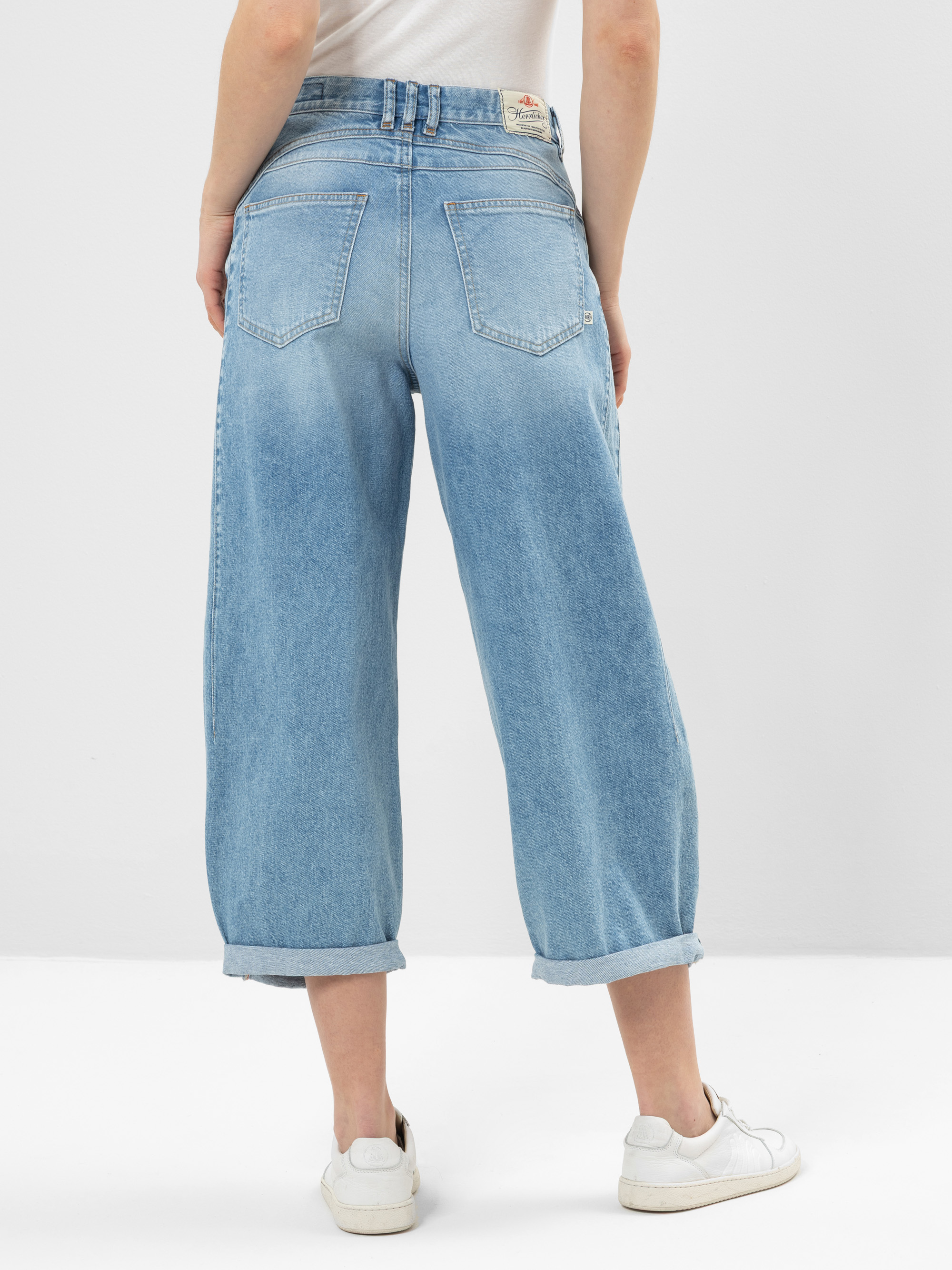 Person in hellblauer, weit geschnittener Jeans mit gekrempelten Bündchen und weißen Turnschuhen, von hinten gesehen. Die Jeans hat zwei große Gesäßtaschen und einen hellbraunen Aufnäher am Bund. Der Hintergrund ist einfarbig und hell gefärbt.