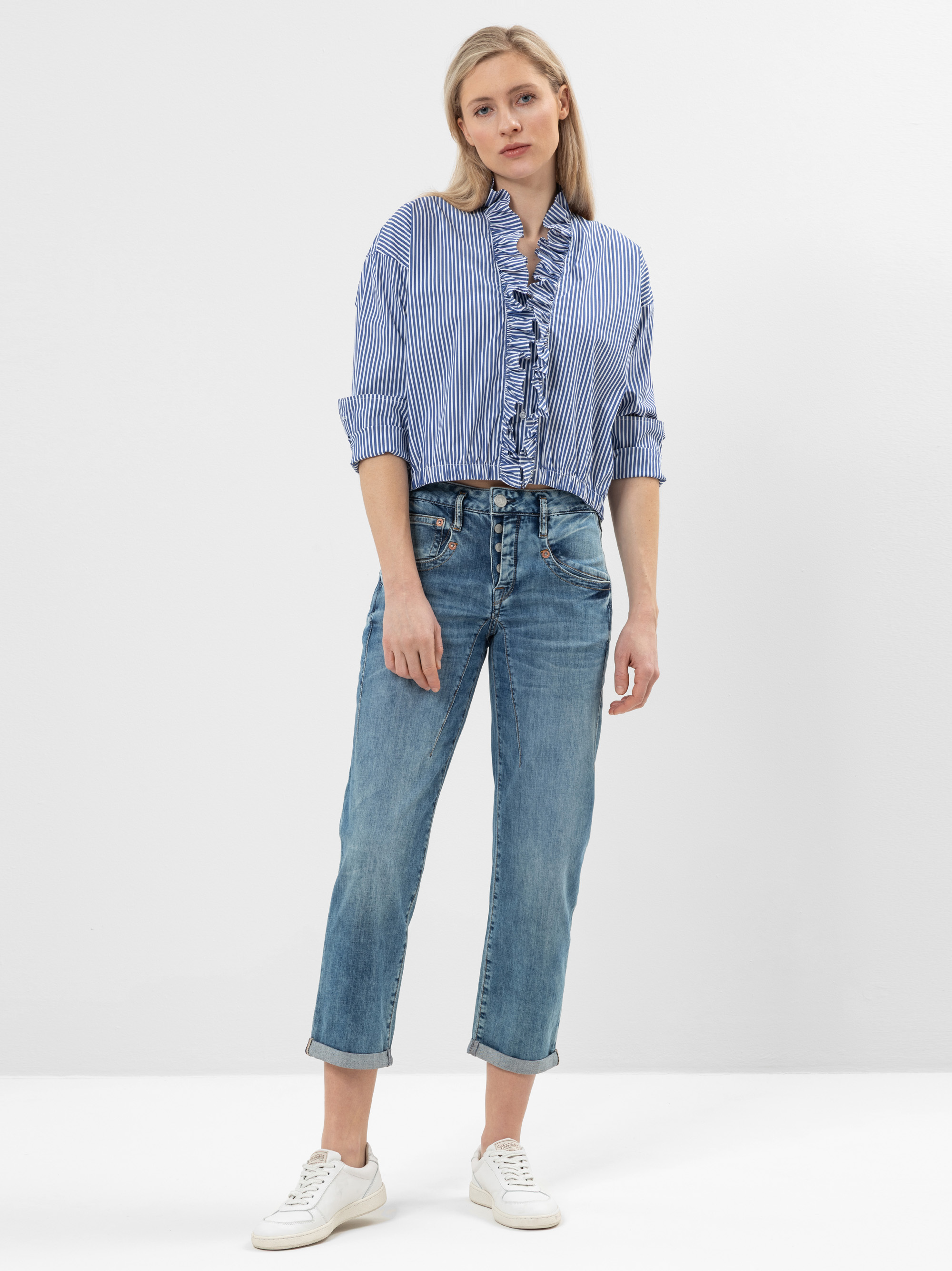 Eine Frau steht vor einem schlichten Hintergrund und trägt eine blau-weiß gestreifte Rüschenbluse, eine kurze Bluejeans und weiße Turnschuhe. Sie hat helles Haar und einen neutralen Gesichtsausdruck, ihre Arme liegen entspannt an ihren Seiten.