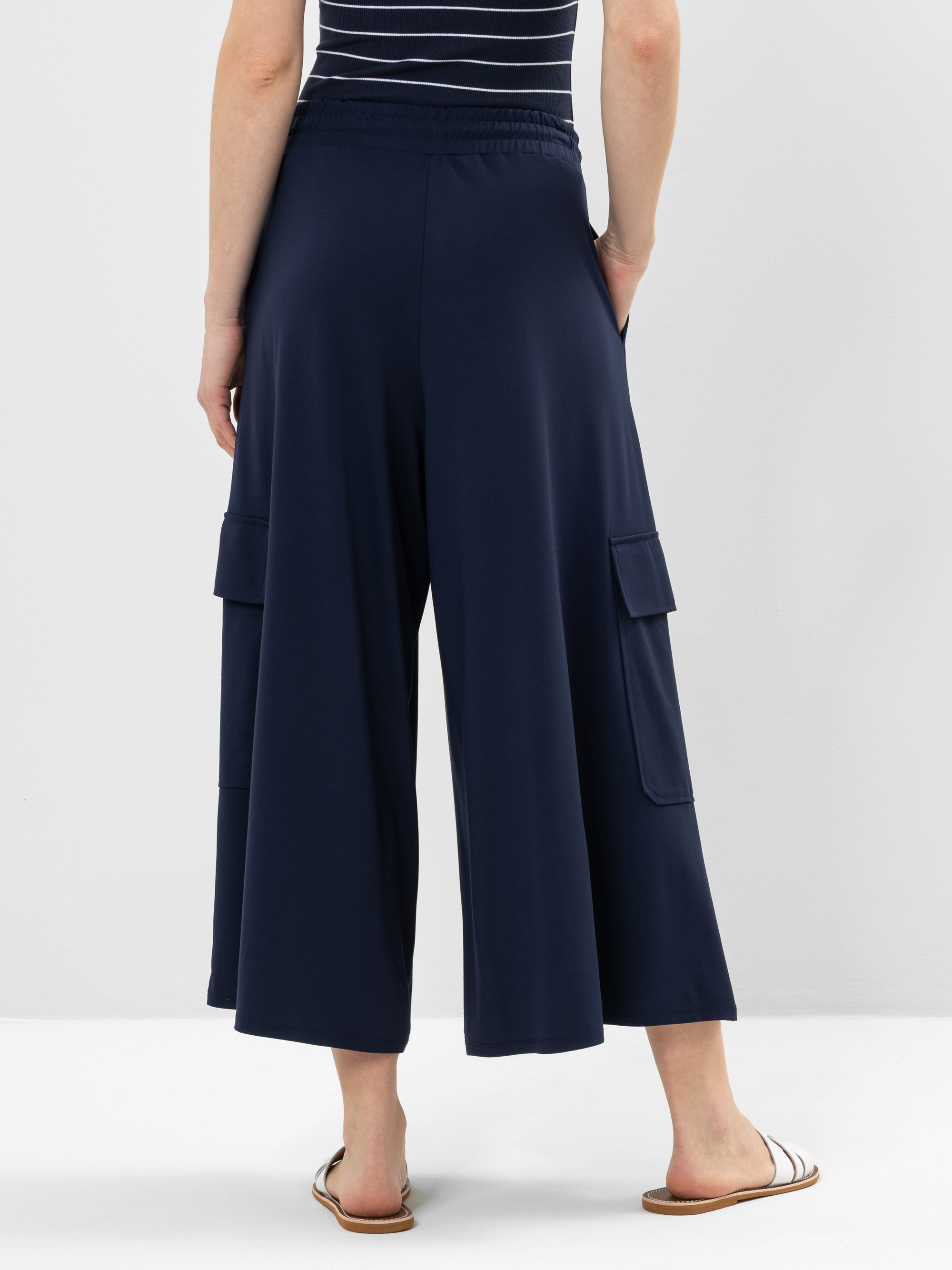 Eine Person, die eine marineblaue, weit geschnittene Hose mit seitlichen Cargotaschen, ein schwarz-weiß gestreiftes Oberteil und weiße Sandalen trägt, steht vor einem schlichten, hellen Hintergrund.