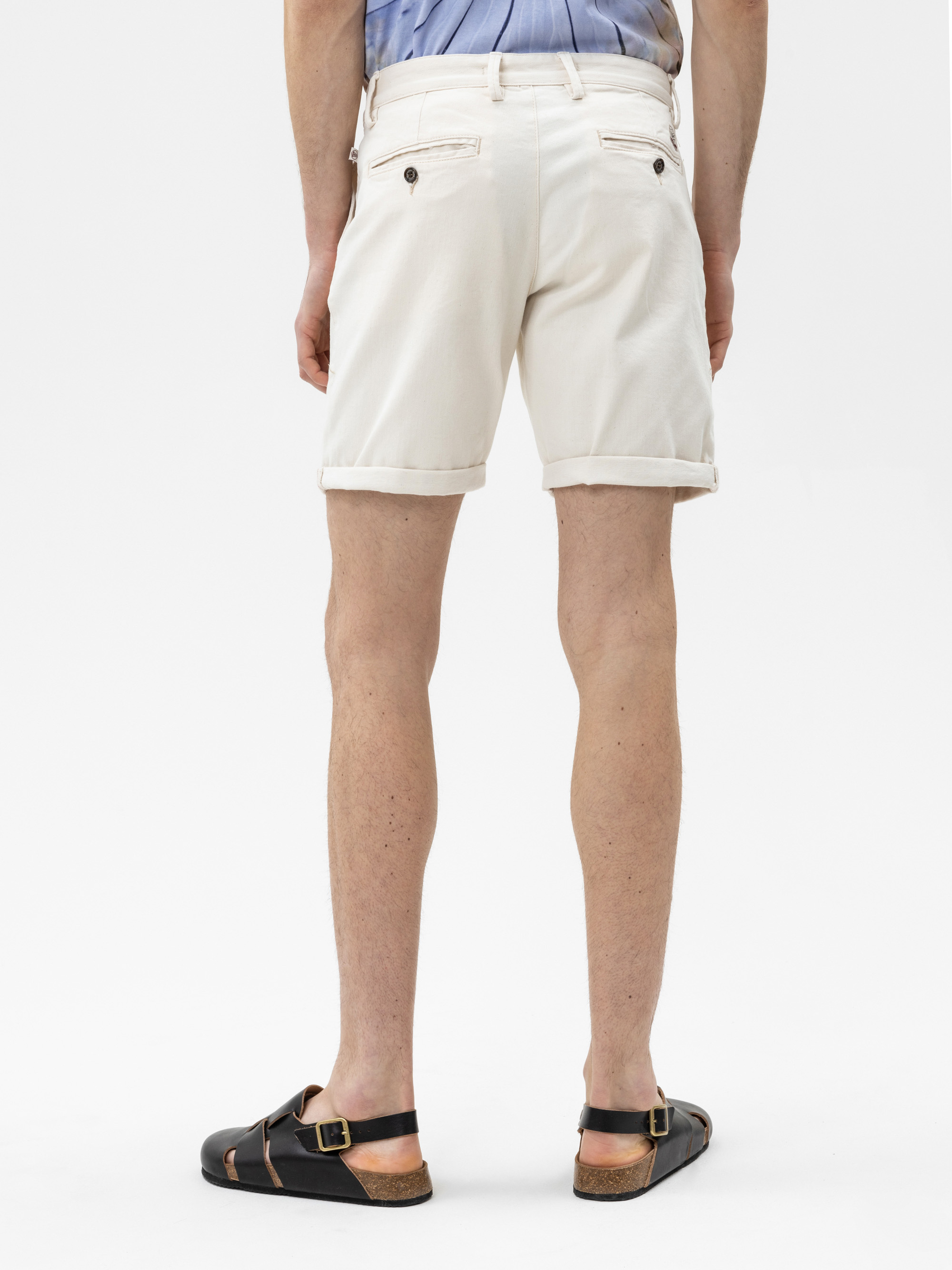Eine Person, die cremefarbene Shorts mit Bündchen, ein hellblaues Hemd und schwarze Sandalen trägt, ist von hinten zu sehen und steht vor einem schlichten weißen Hintergrund.